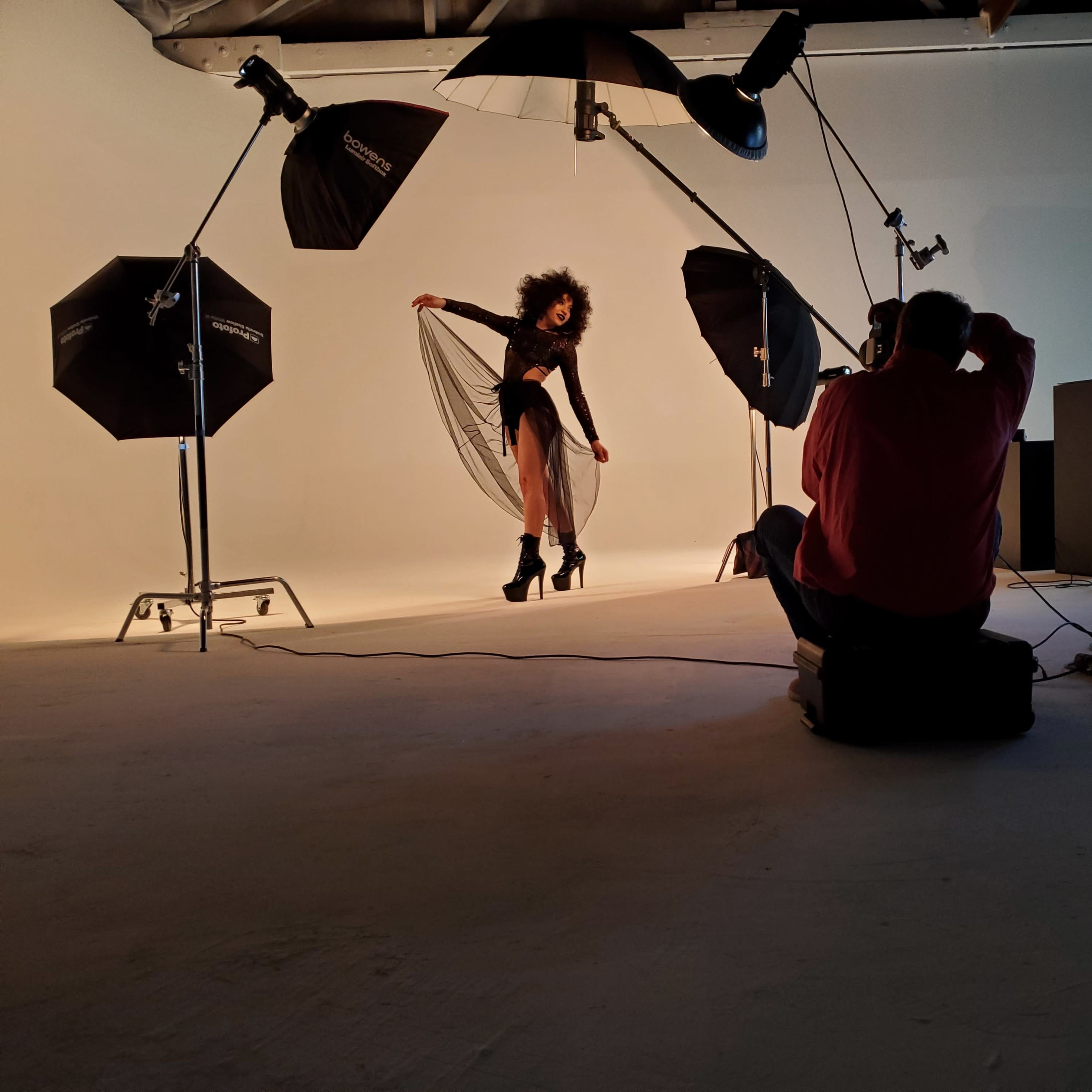 A model poses with a flowing dress on a photoshoot set, while a photographer reviews images on a computer screen.