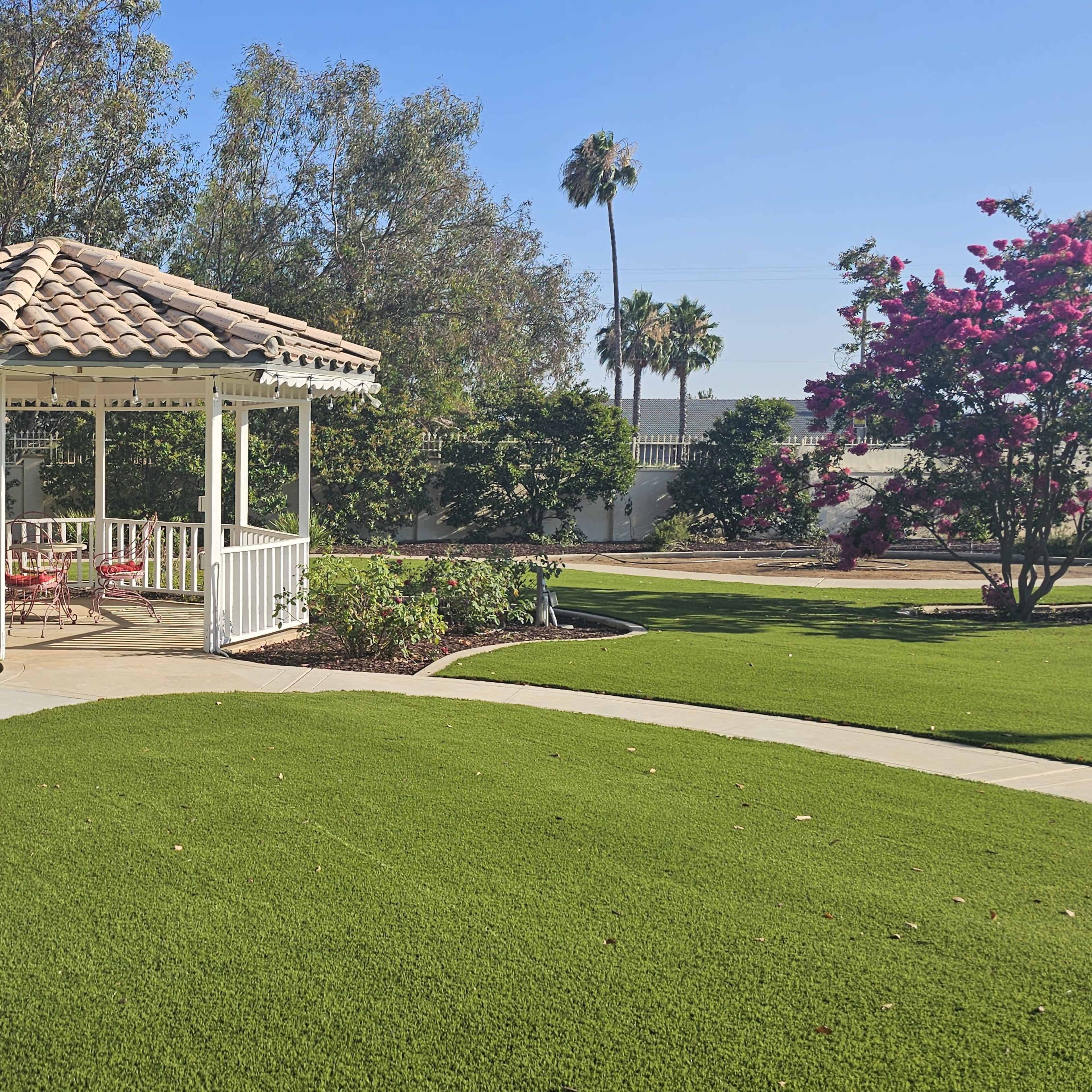A white gazebo with a tiled roof is situated in a landscaped garden featuring green grass and flowering shrubs.