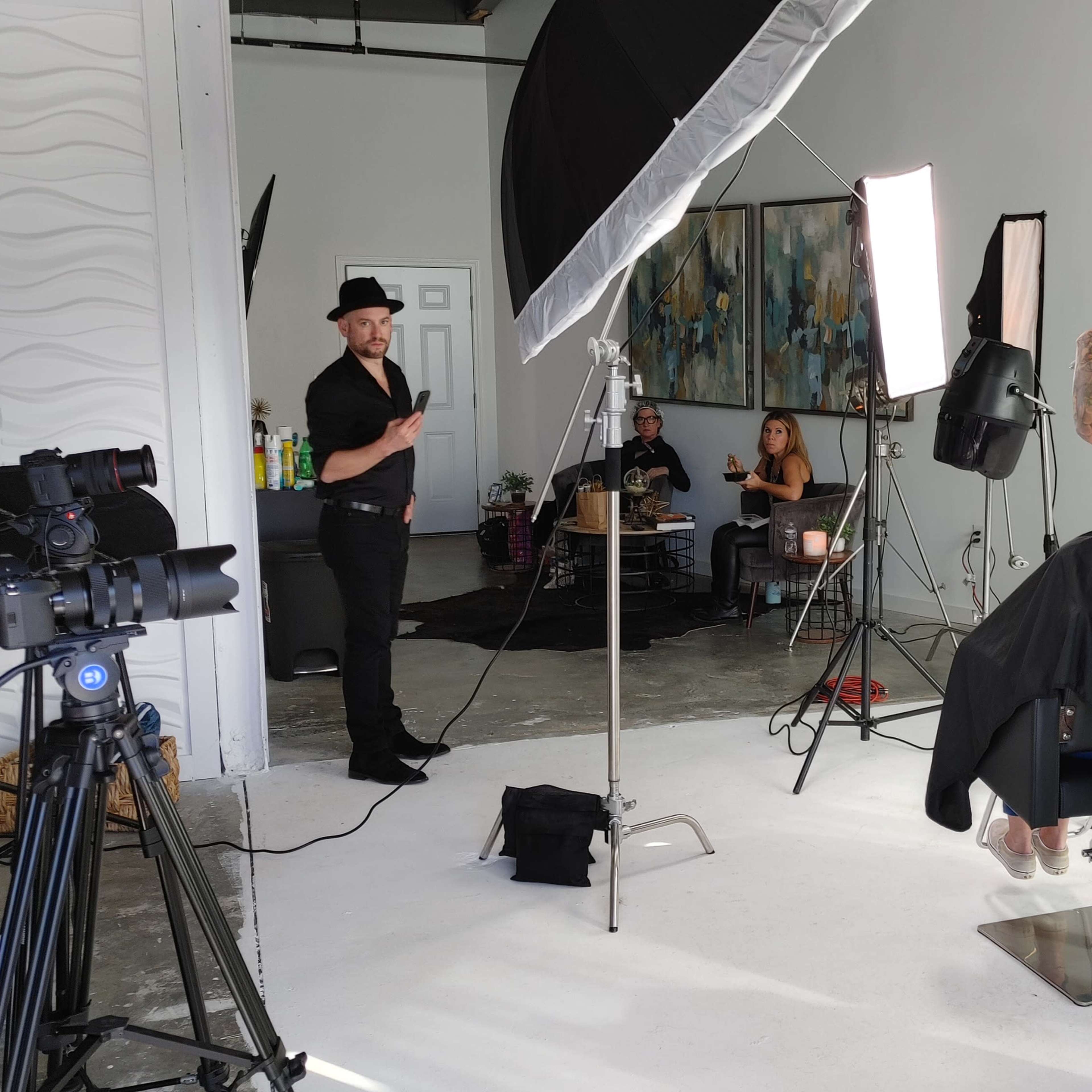 A hair stylist works on a client's hair in a studio setting, while a photographer and two models observe in the background.