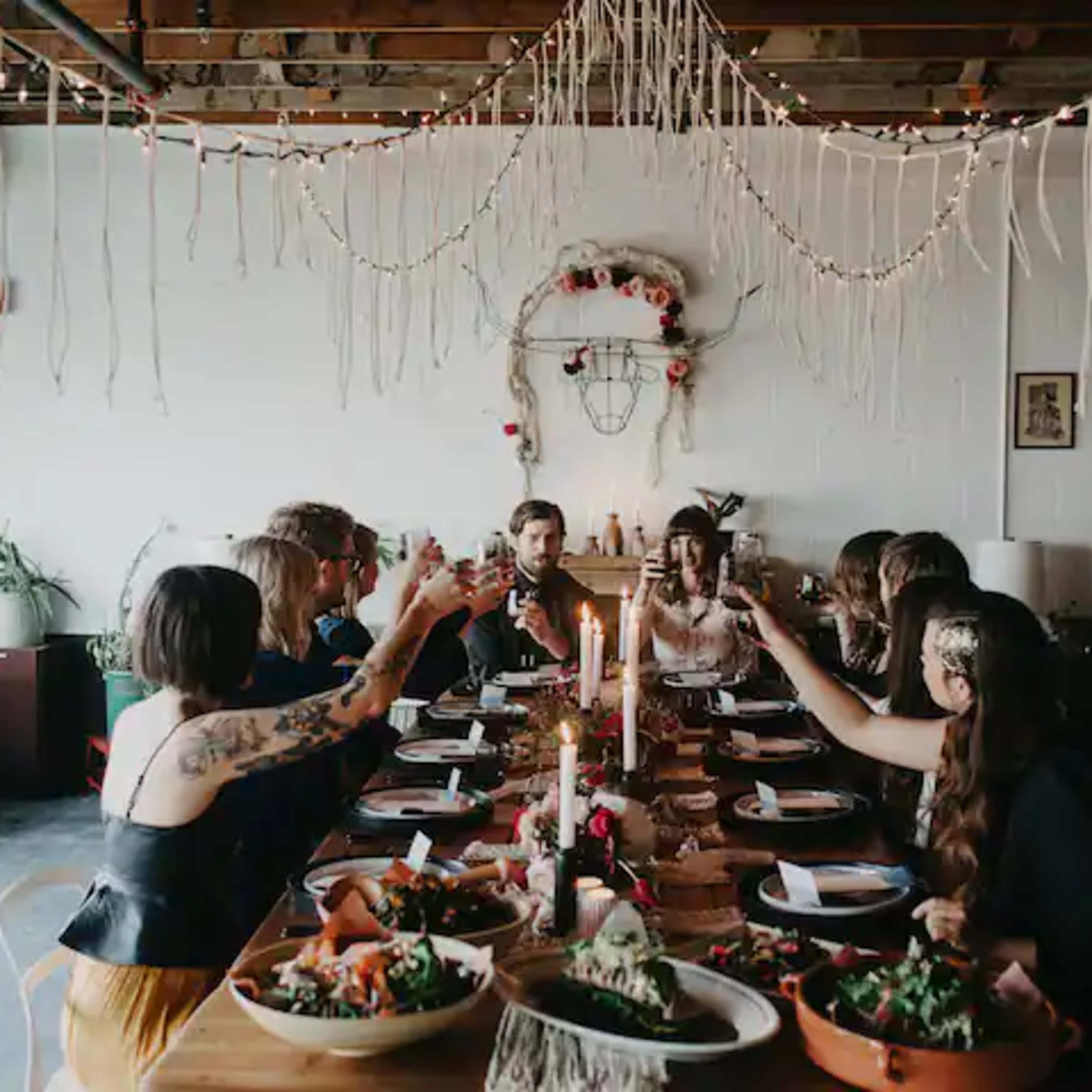 A group of people sits around a long table decorated with candles and food, raising their glasses in a toast.