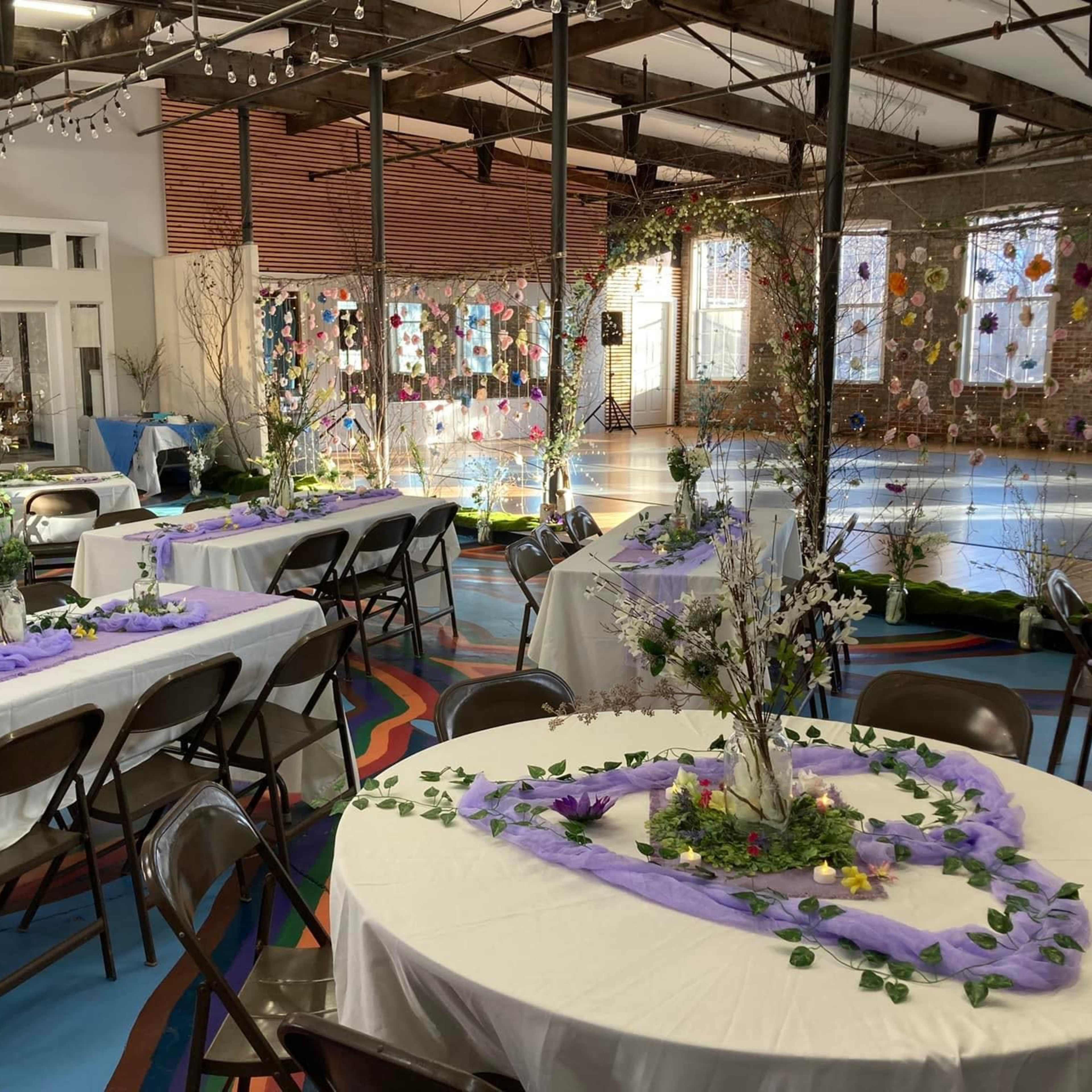 A spacious event hall features several round tables adorned with floral centerpieces, surrounded by chairs and decorated with fabric drapes, while colorful artwork adorns the walls.