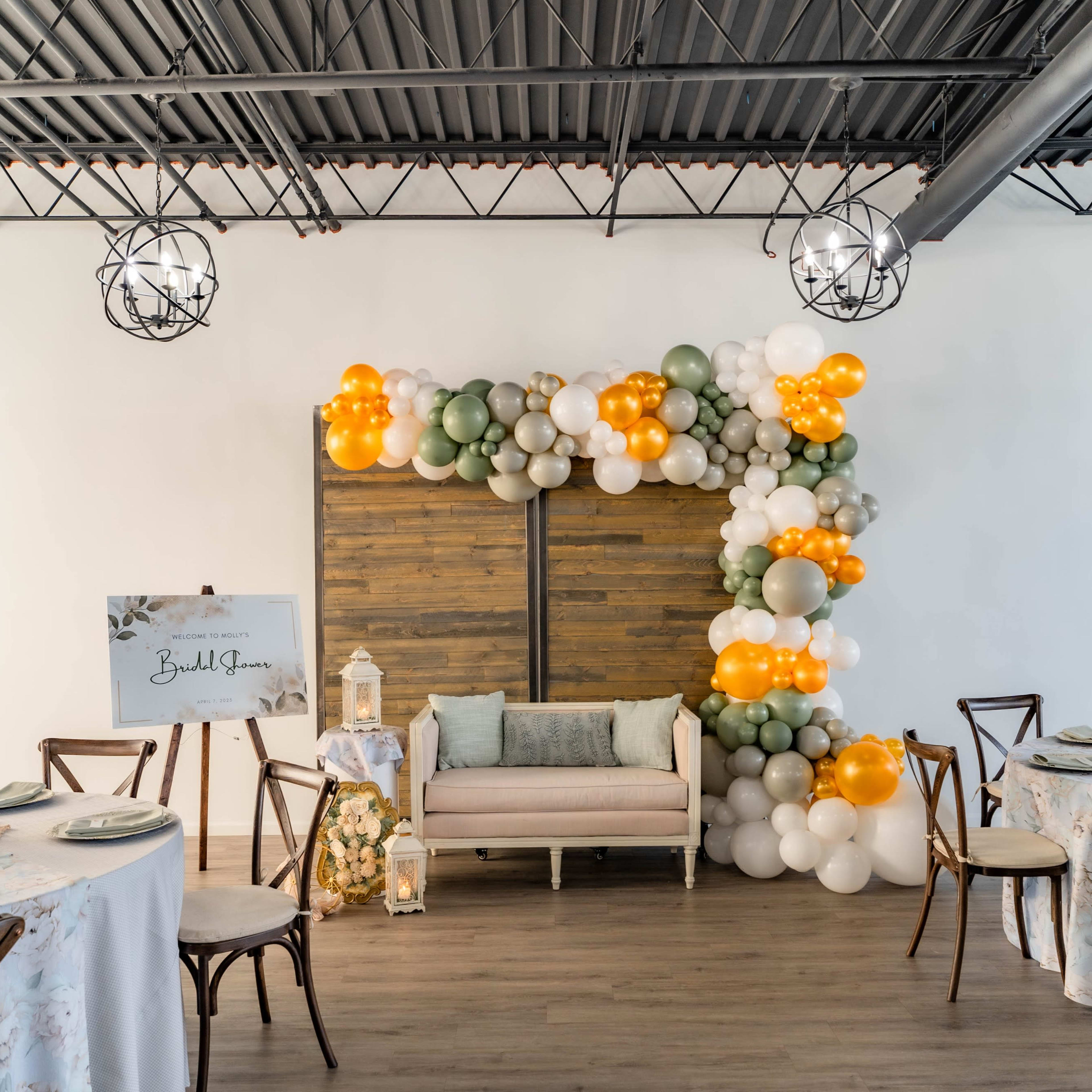 A decorated event space with balloon arrangements in orange, green, and white, a wooden backdrop, and elegantly set tables.