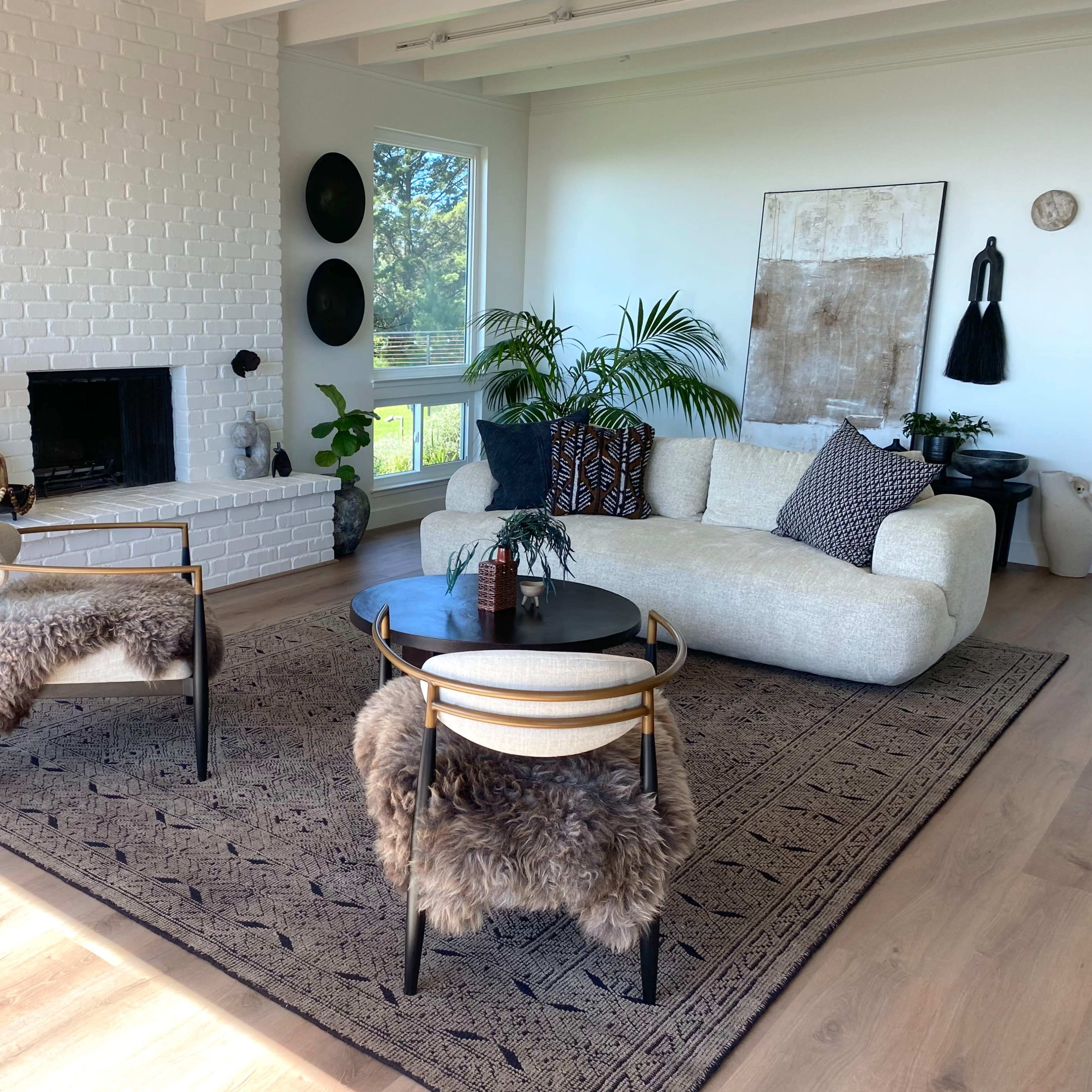 A modern living room with a white brick fireplace, two chairs with fur throws, a curved sofa, and large windows allowing natural light to fill the space.