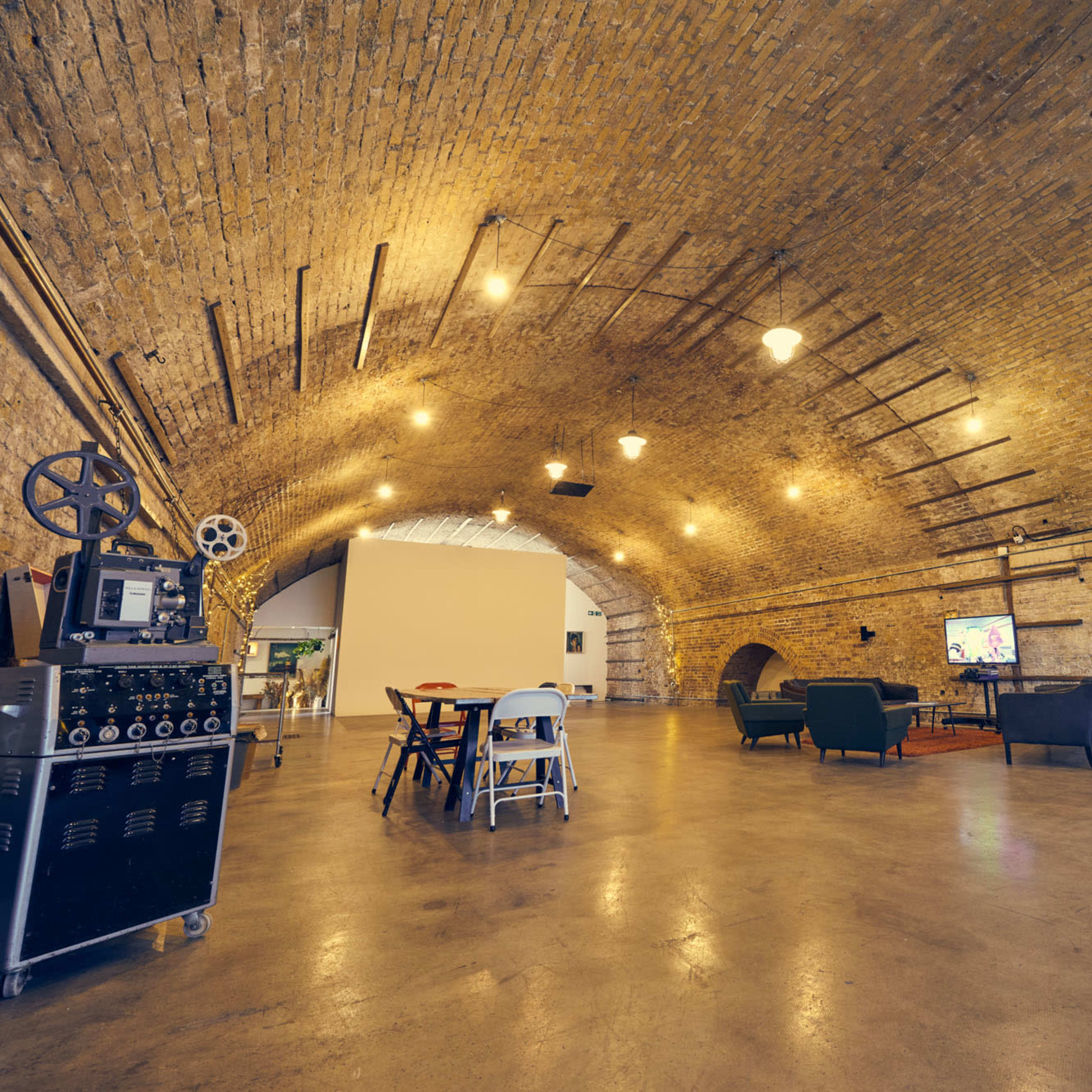 A spacious room with an arched brick ceiling, featuring industrial lighting, wooden furniture, and a decorative TV area.