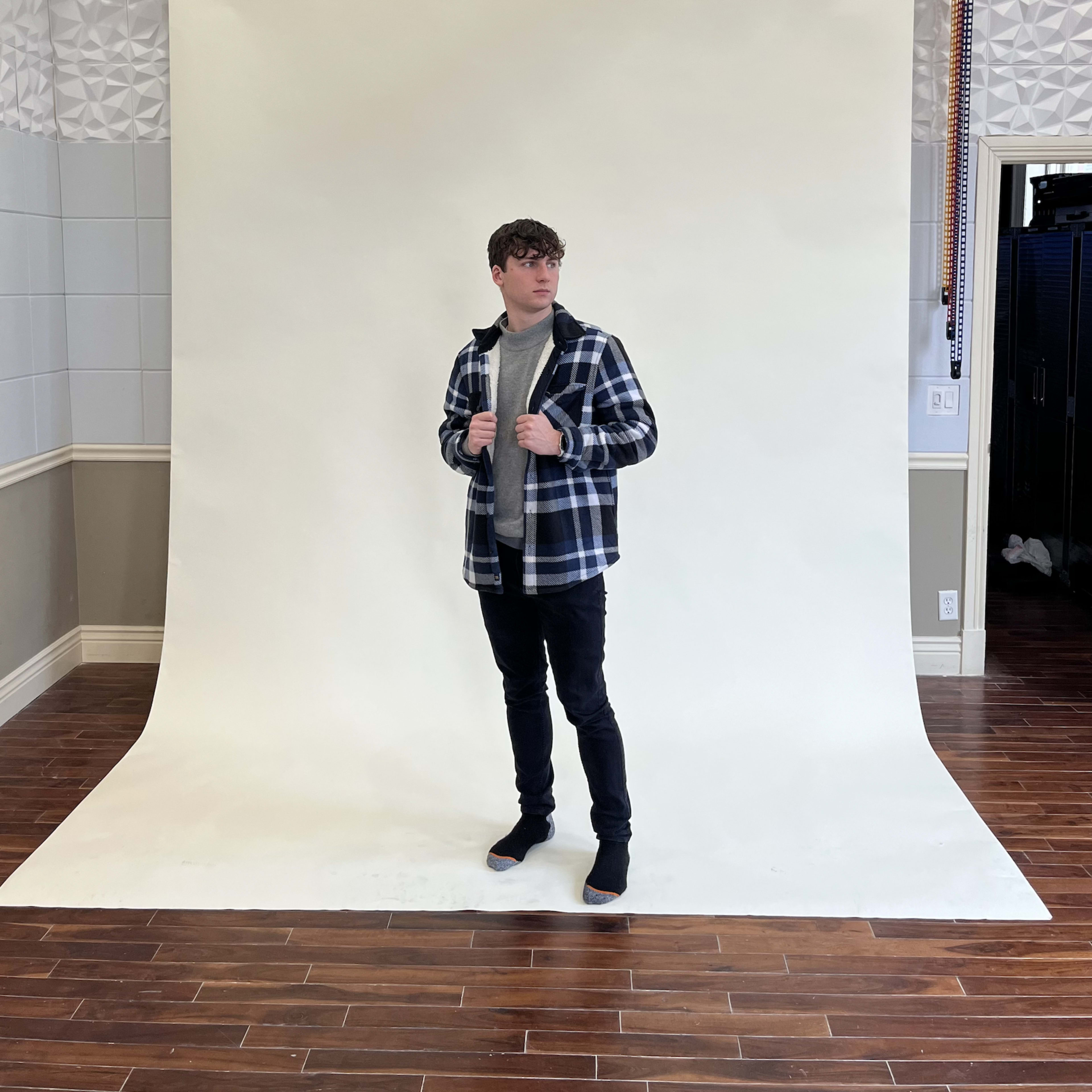 A person in a checkered jacket poses in front of a plain white backdrop in a studio.