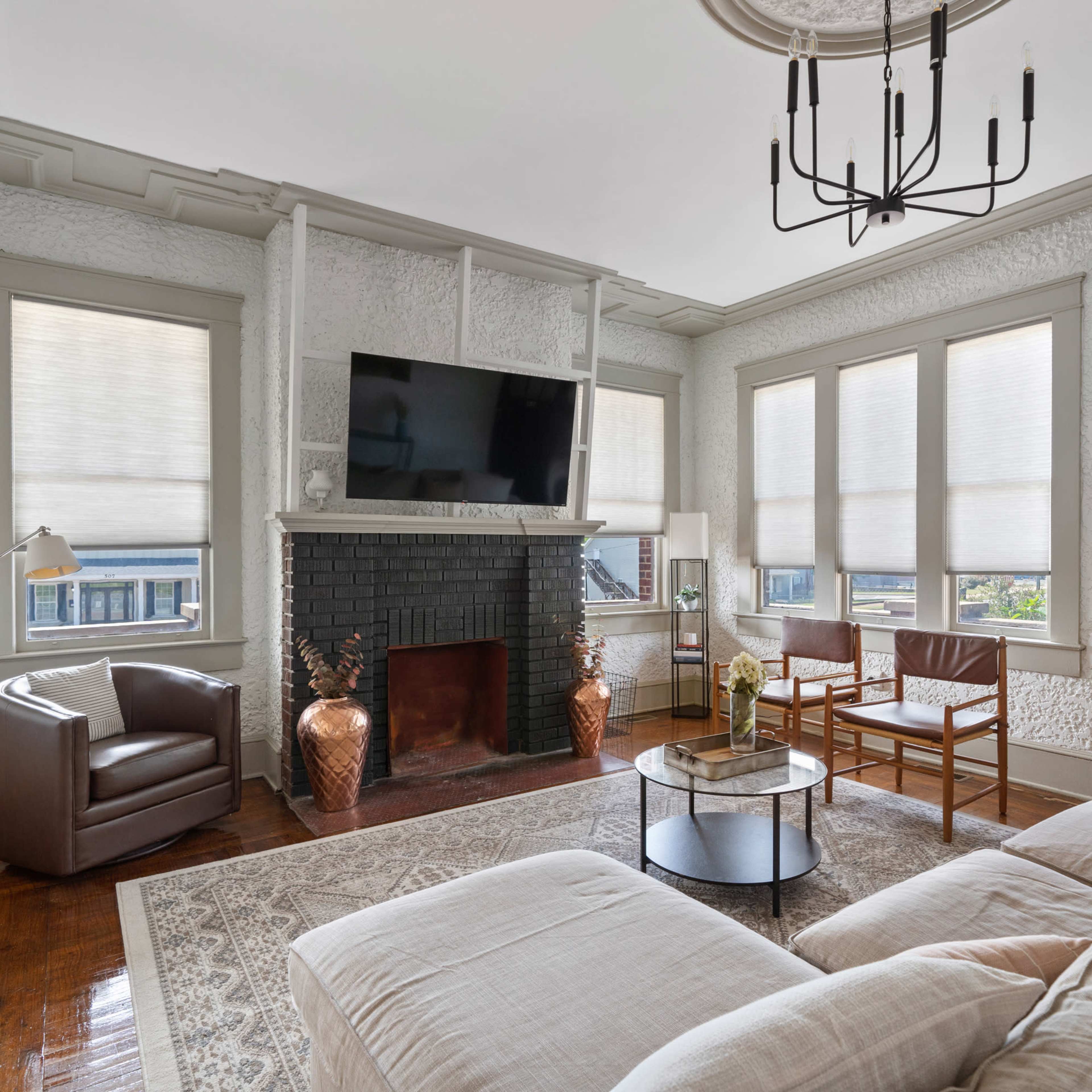 The living room features a sectional sofa, a coffee table, a mounted television over a black fireplace, and large windows with shades.