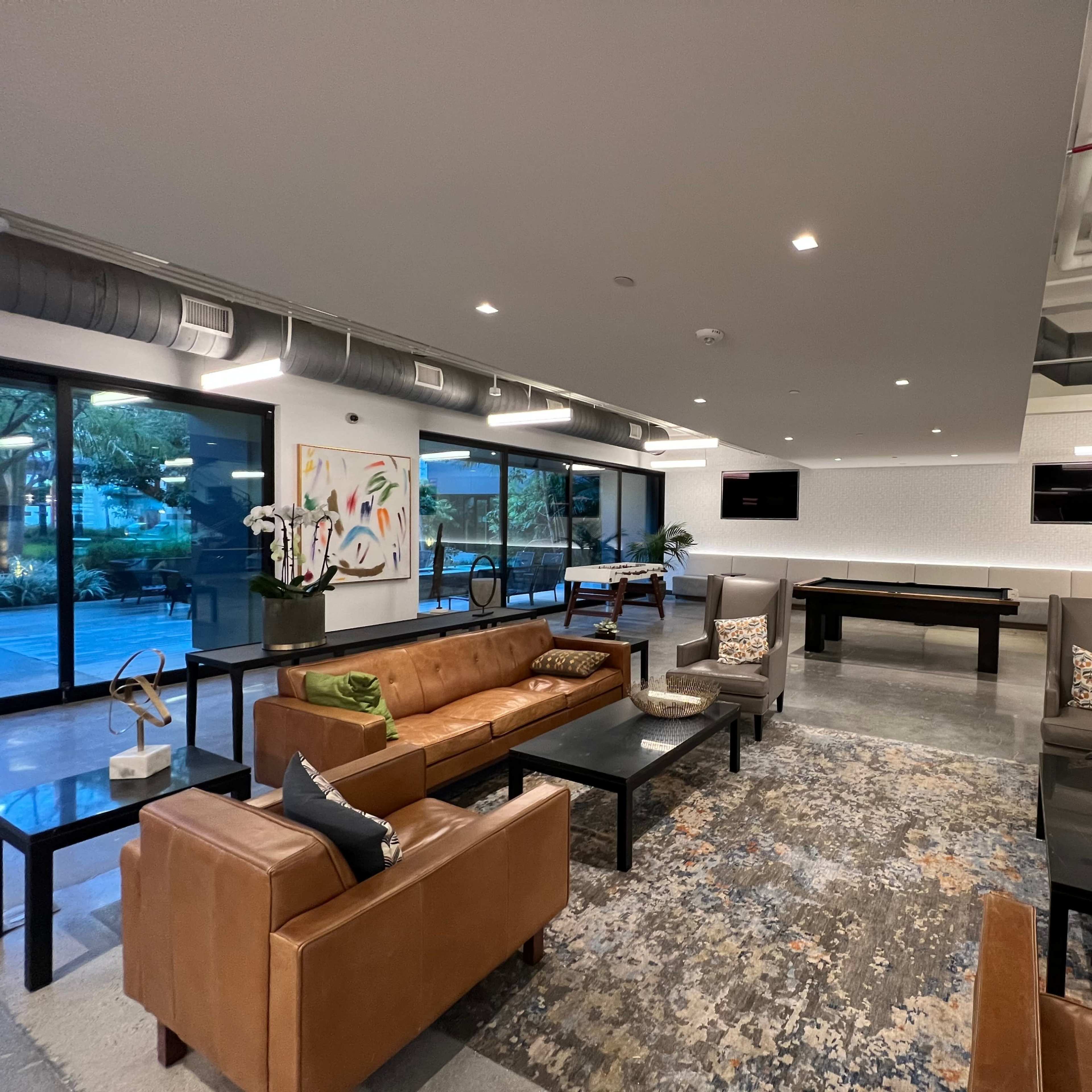 A contemporary lounge area featuring leather seating, a coffee table, and a billiards table, all surrounded by large windows.