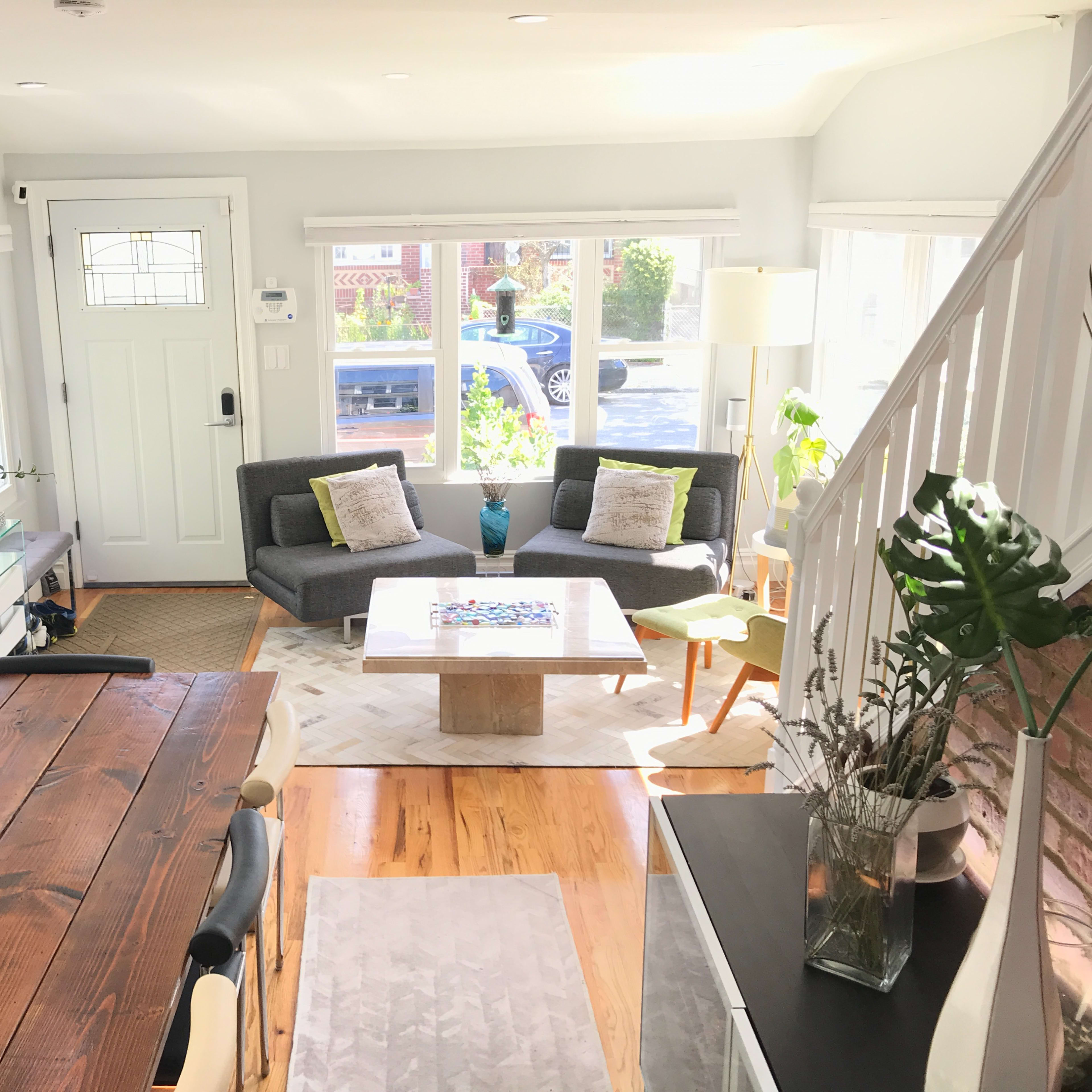 A bright living space featuring a gray sofa, a coffee table, a dining area with a wooden table, and large windows that allow natural light to enter.