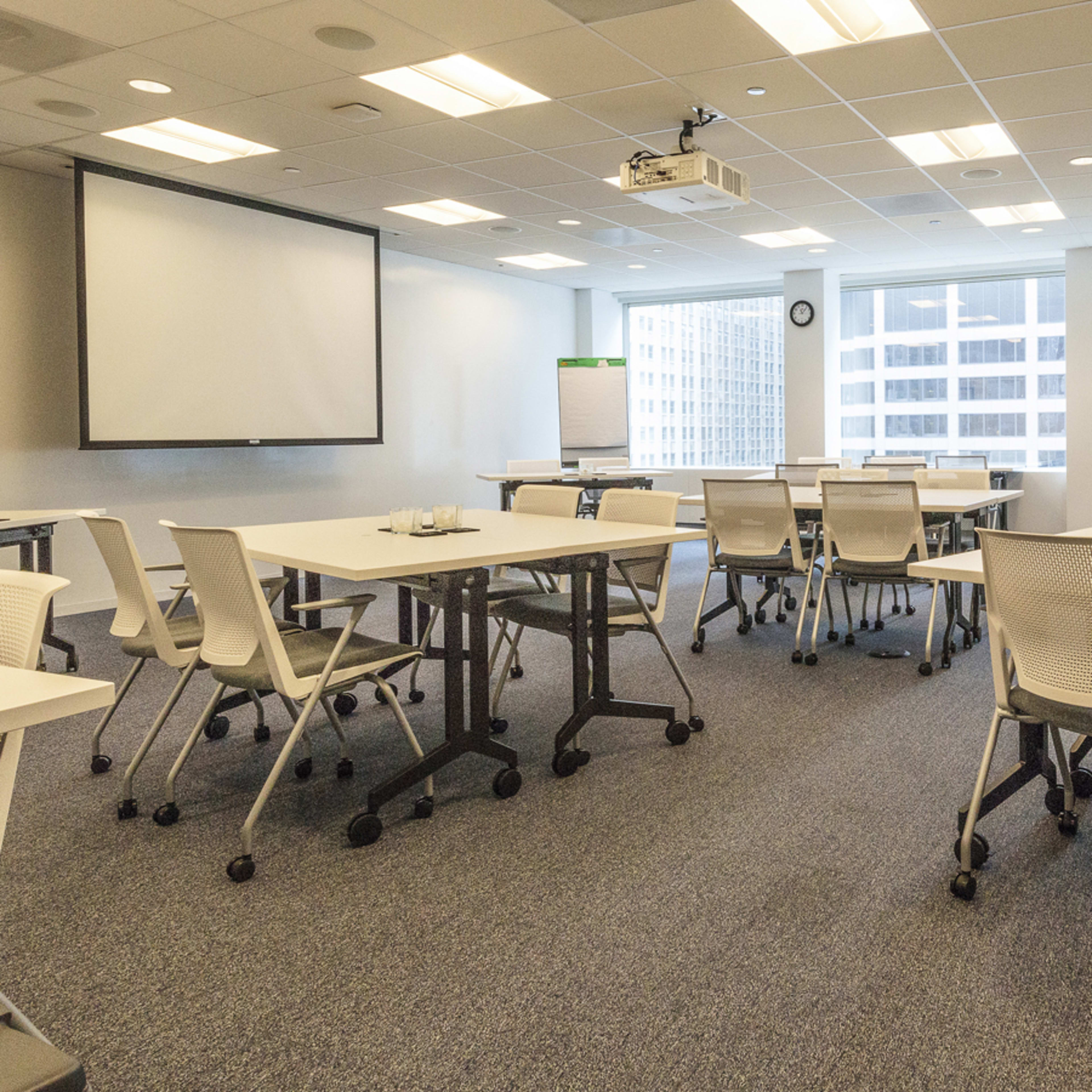 A modern conference room with several movable tables and chairs arranged around a projection screen and large windows.