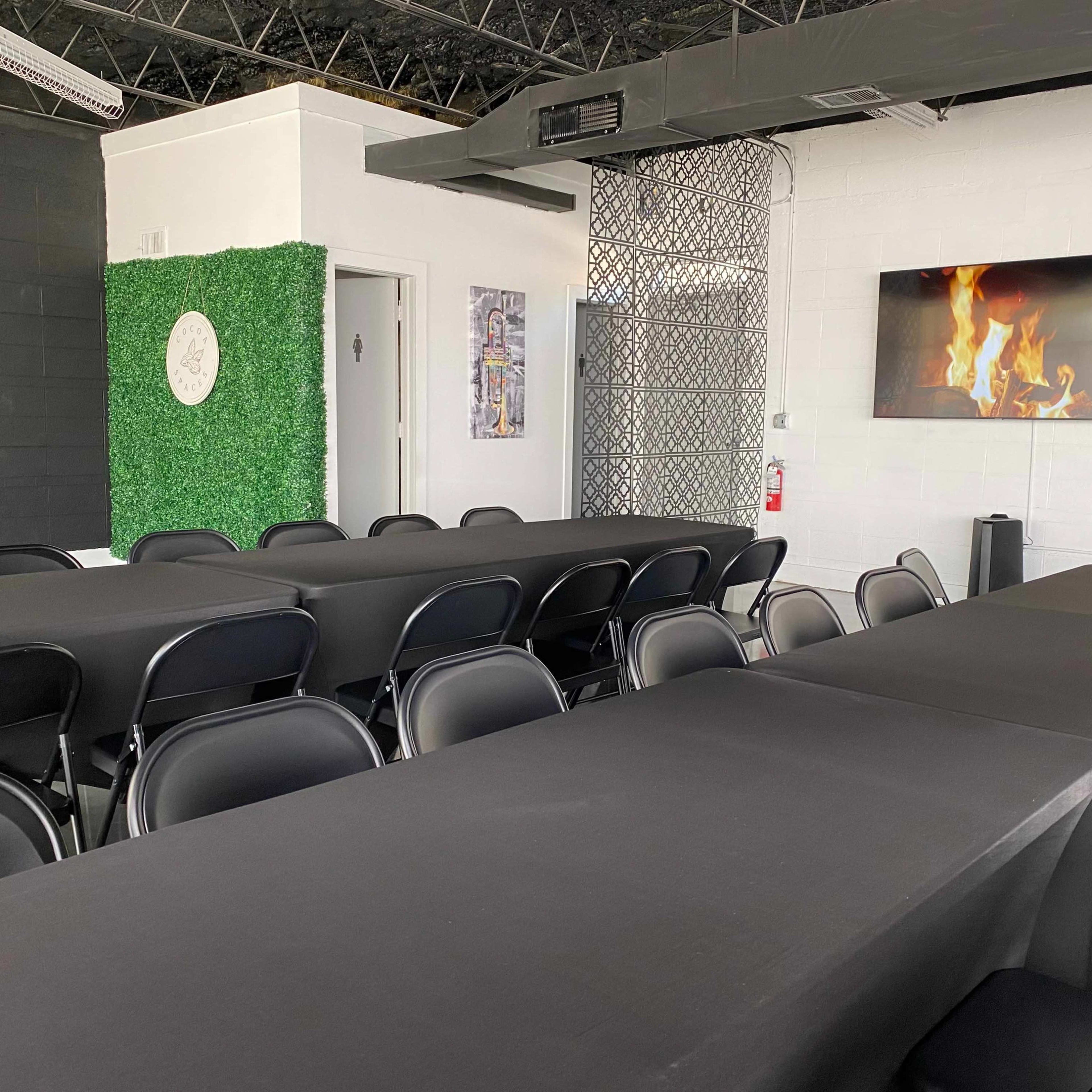 A modern conference room features long black tables with black chairs, a green wall with decorative elements, and a screen displaying a fireplace image.