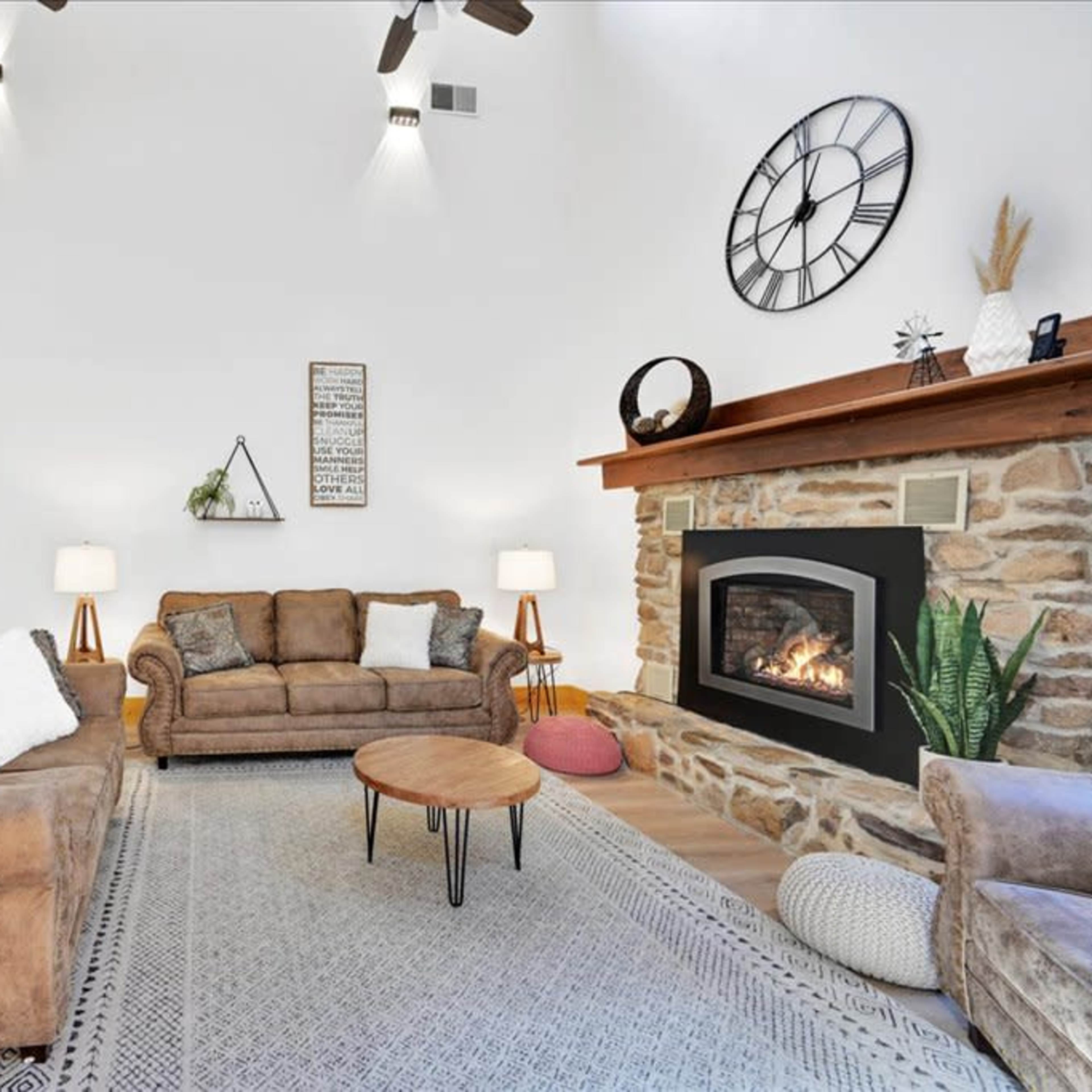 A living room with two brown sofas, a round coffee table, and a stone fireplace beneath a large clock on the wall.