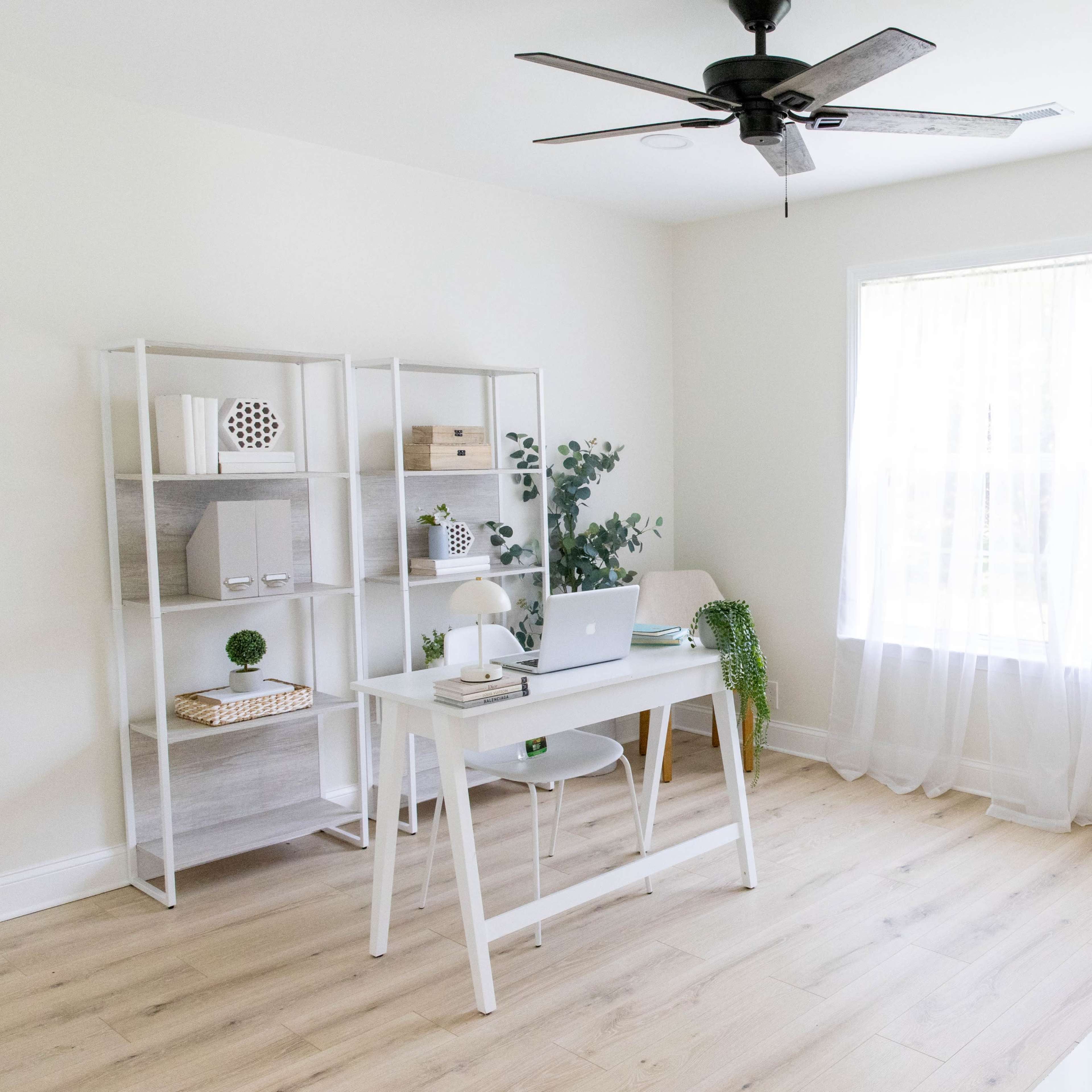 A bright and minimalist home office features a desk with a laptop, a bookshelf filled with decorative items, and a window draped with sheer curtains.