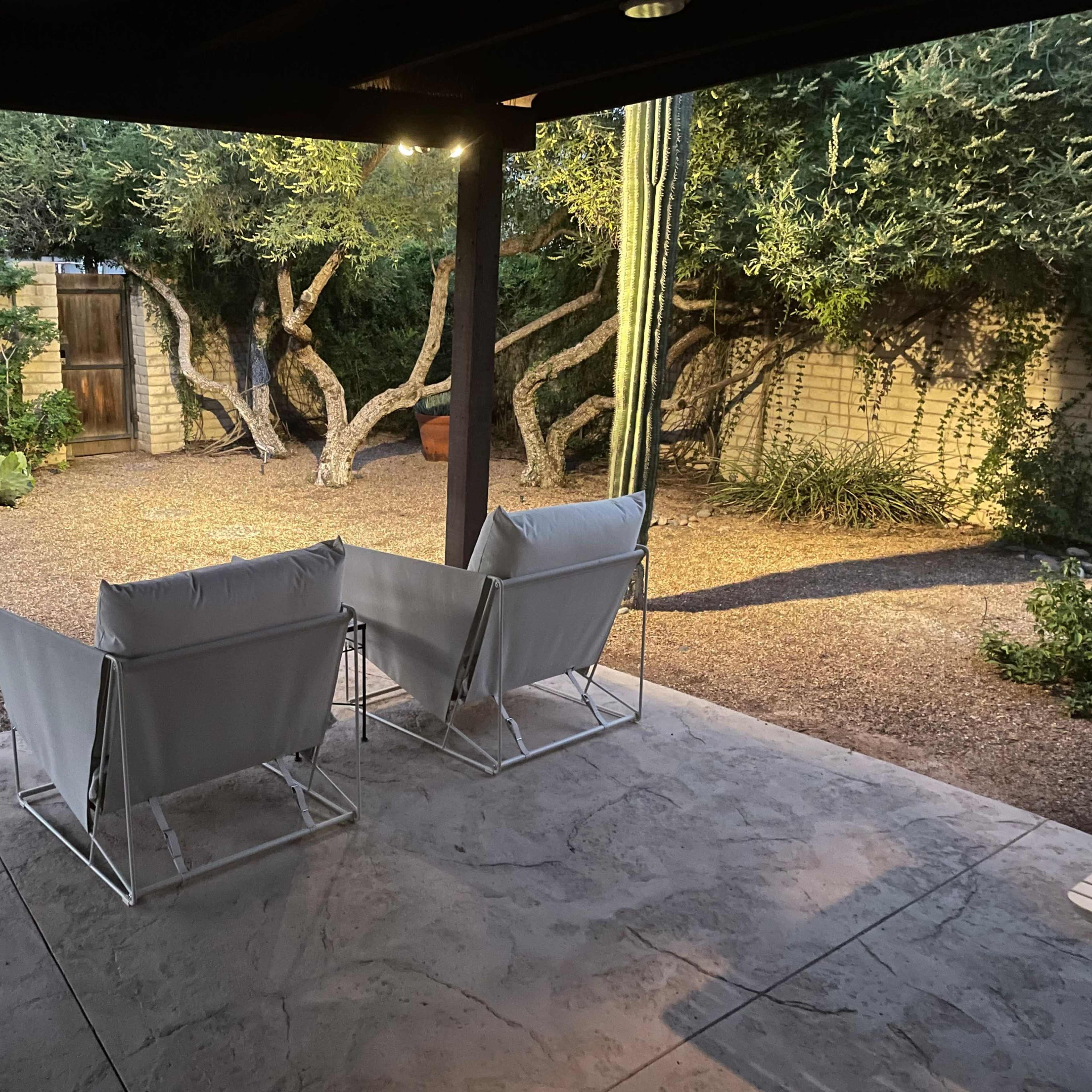 Two white folding chairs are positioned in a shaded patio area, overlooking a gravel garden with various plants and a tall cactus.