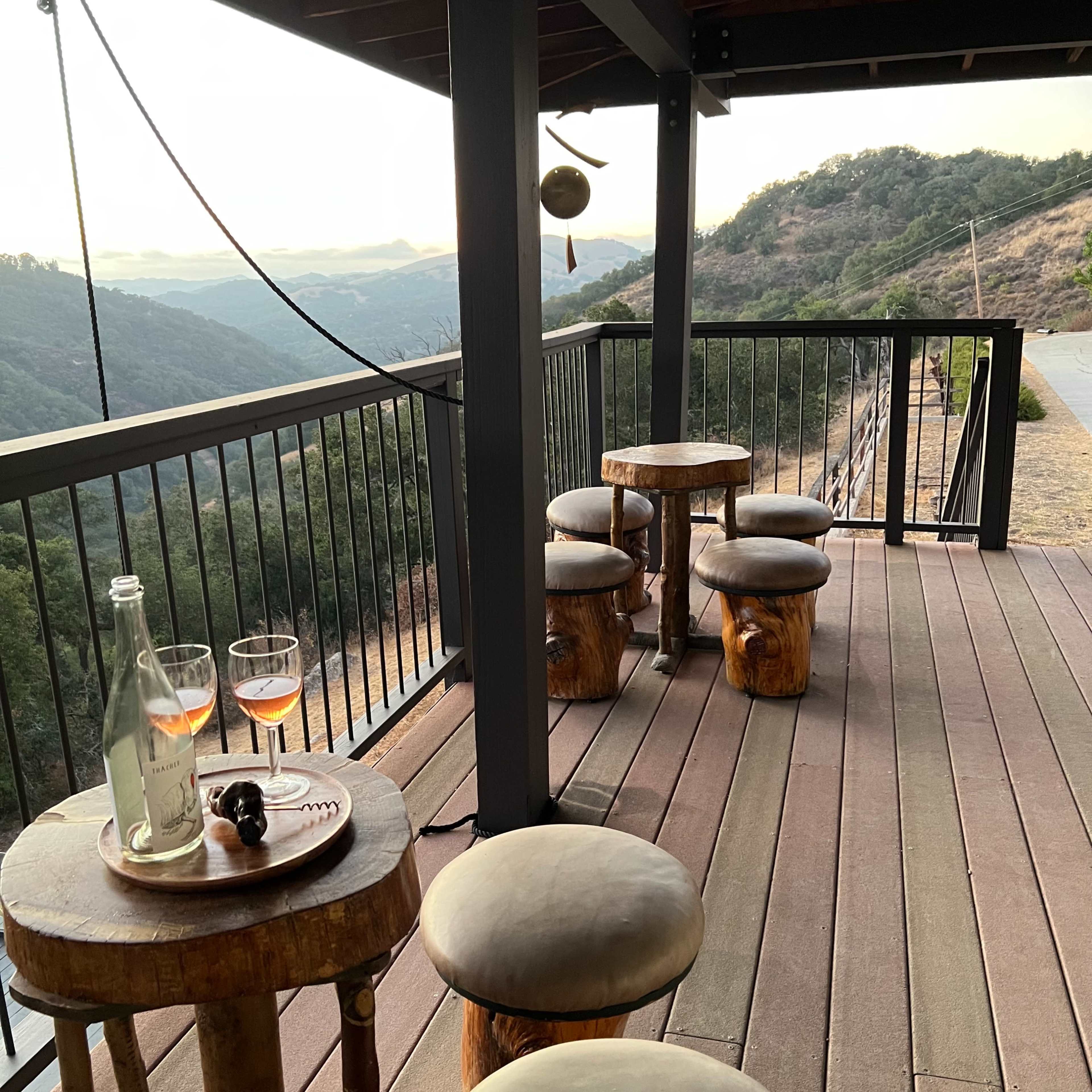 A wooden deck with two glasses of drink and a bottle on a low table, surrounded by log stools and overlooking a hilly landscape.