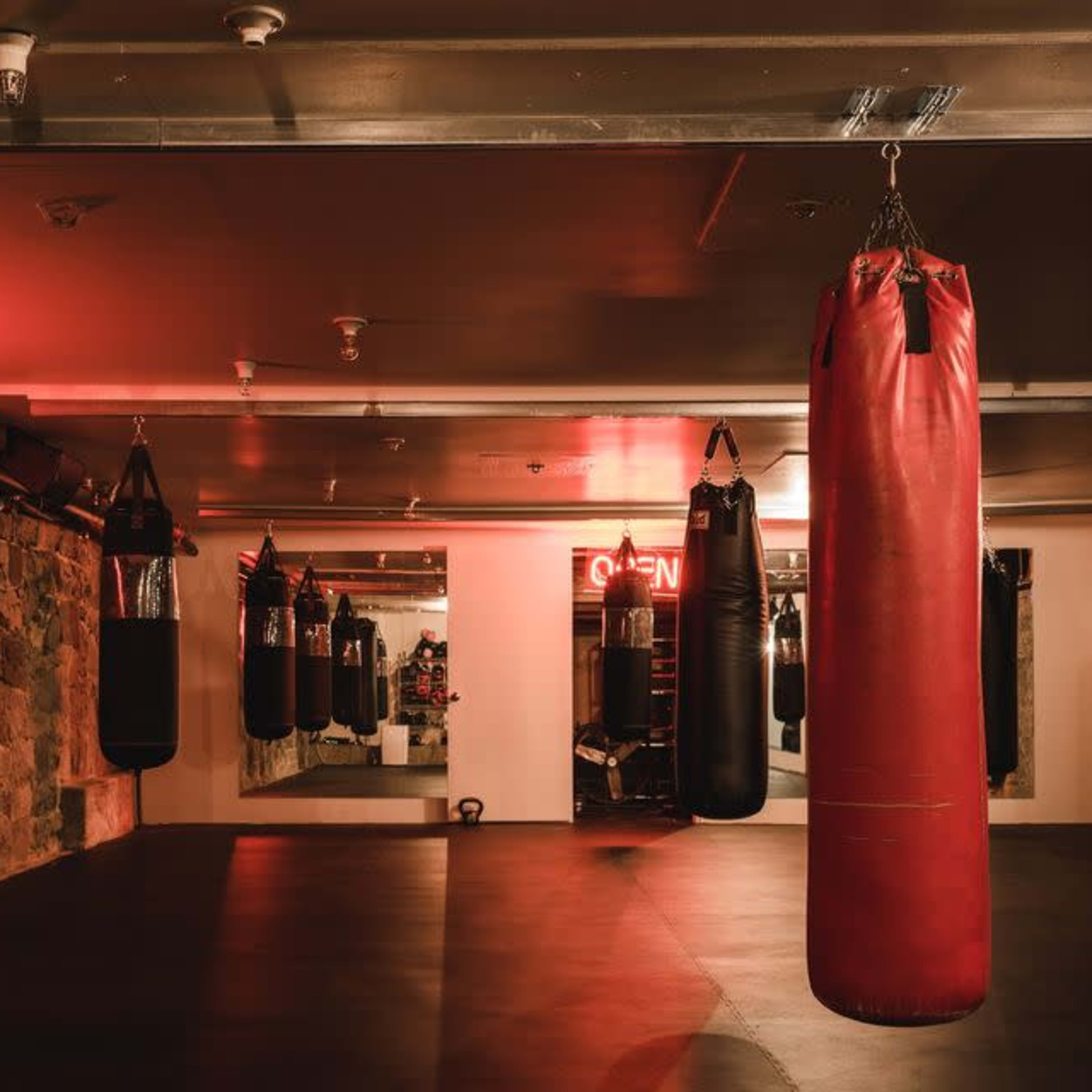 A boxing gym features multiple punching bags hanging from the ceiling, with a neon "OPEN" sign and illuminated walls.