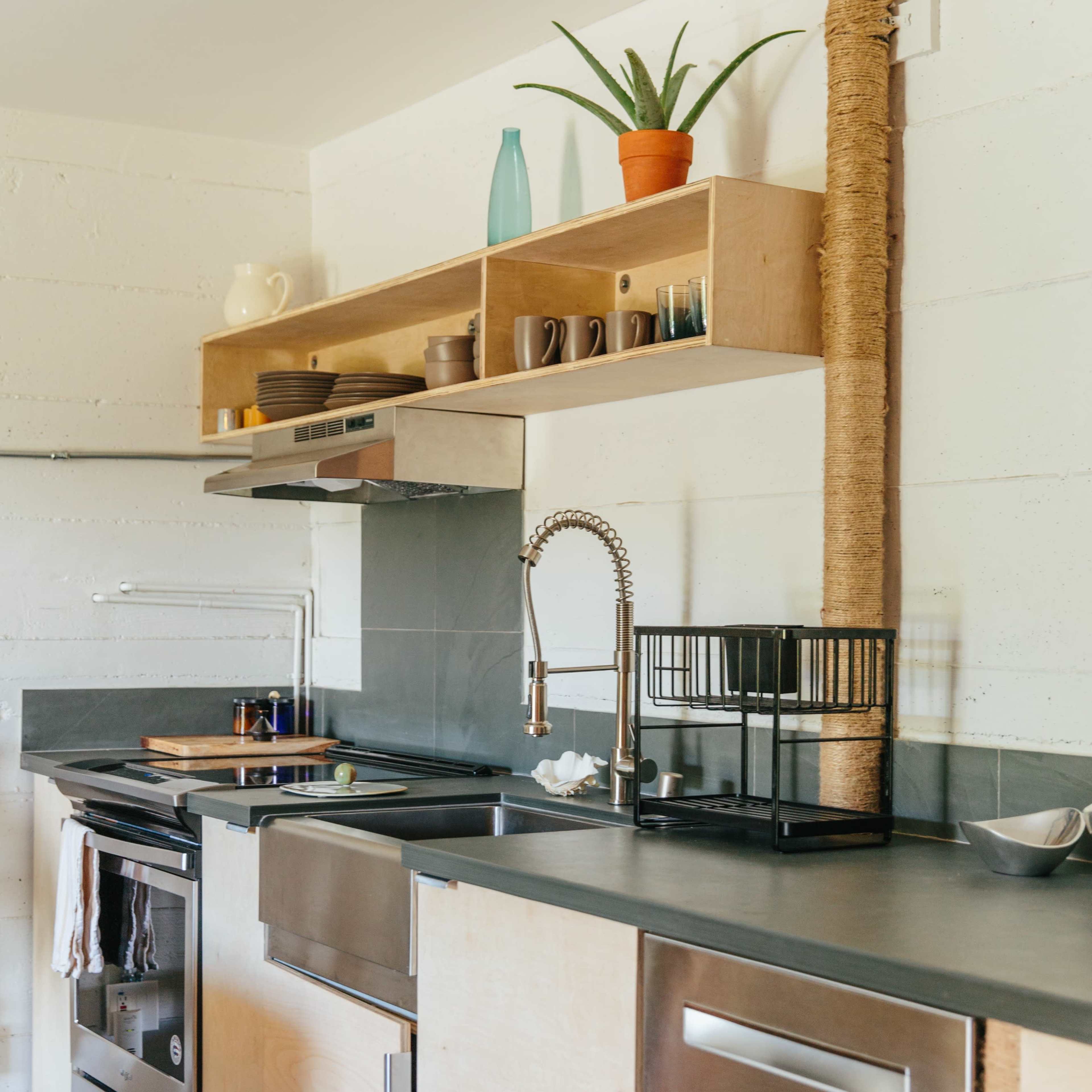 A modern kitchen features a minimalist wooden cabinetry, stainless steel appliances, and a small shelf with potted plants and dishware.