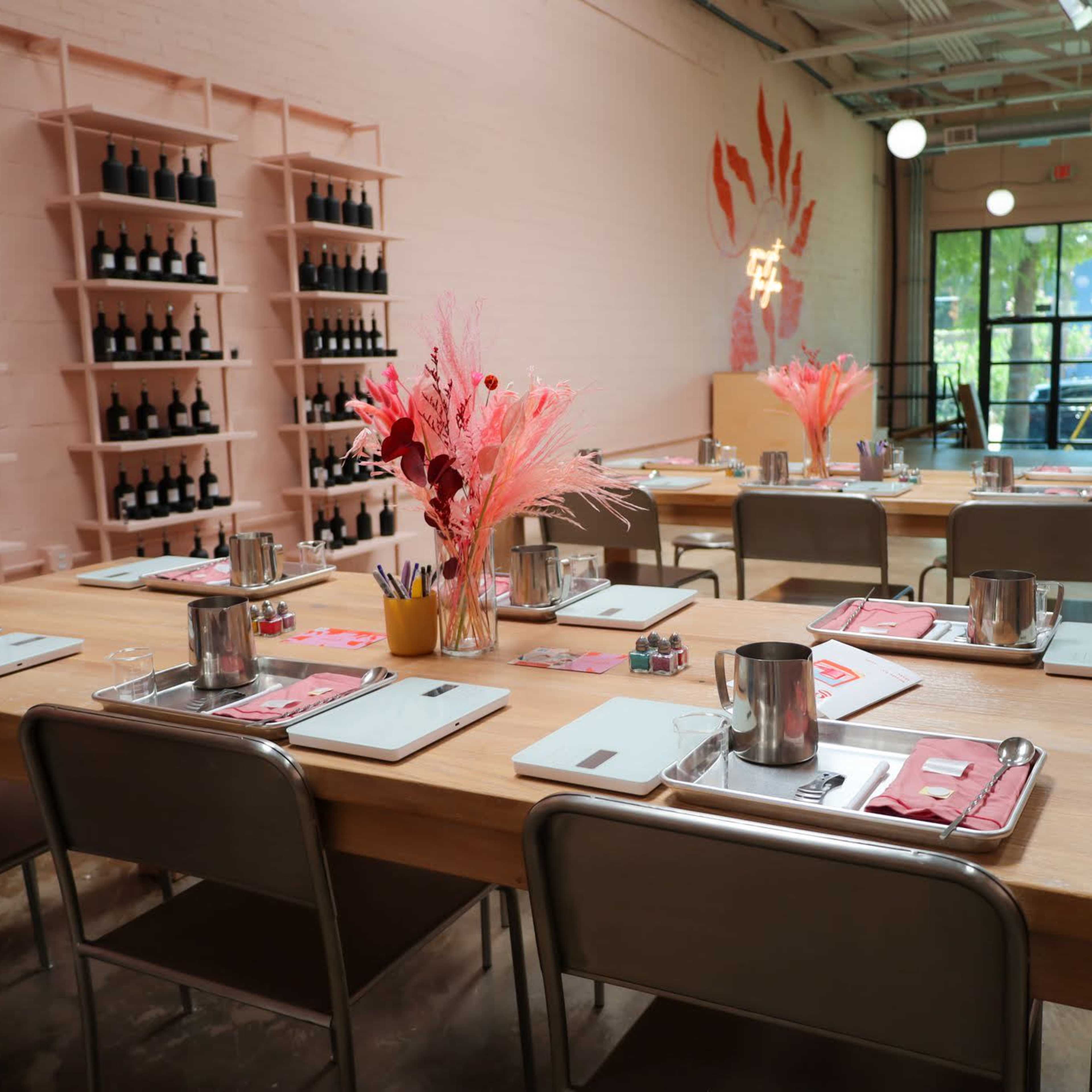 A dining area with wooden tables set for a meal, featuring wine bottles on shelves and decorated with pink floral arrangements.