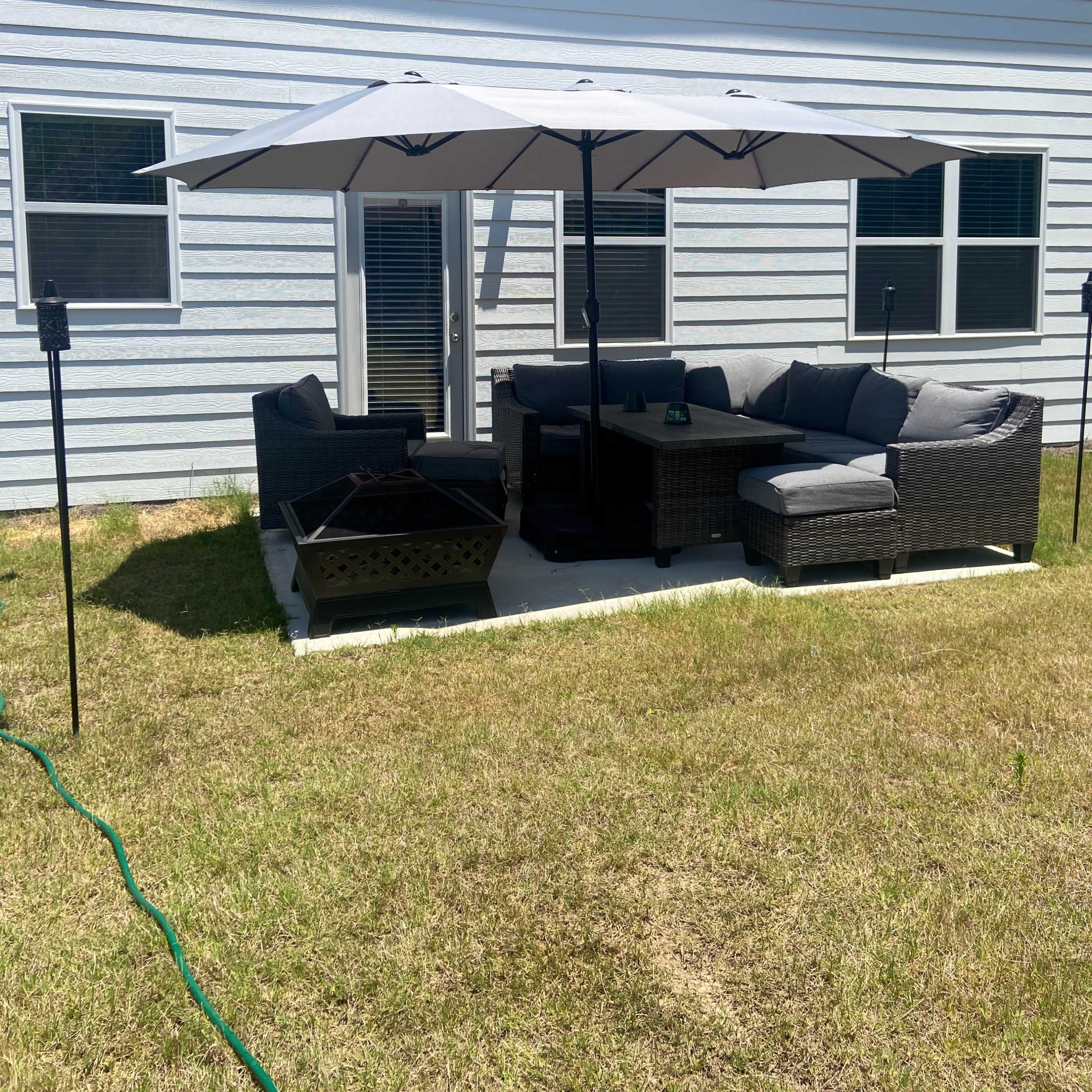 A patio area featuring a sectional sofa and an umbrella is located in a grassy backyard next to a house.