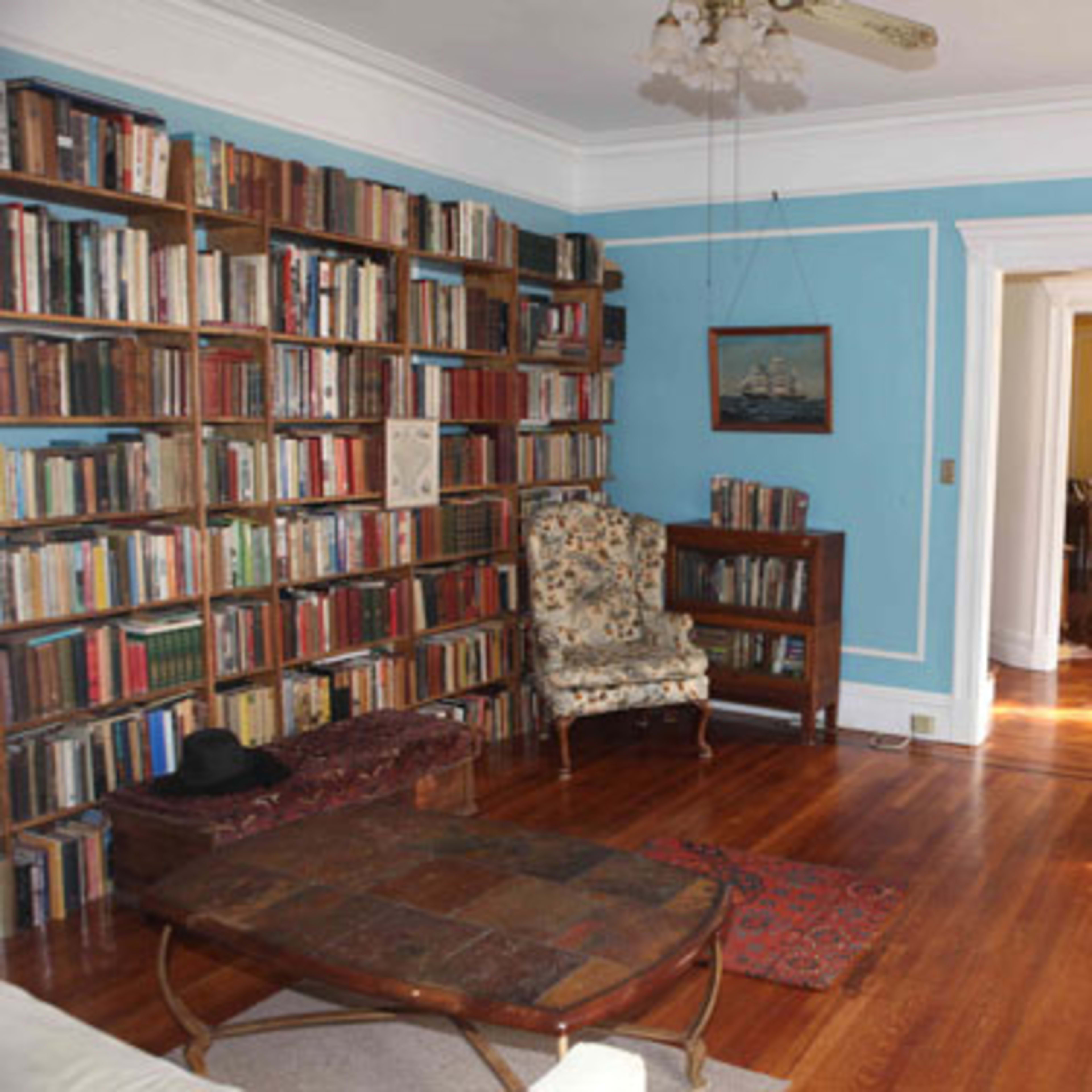 A living room features a blue wall, a large bookshelf filled with books, a patterned armchair, a wooden coffee table, and a TV stand with a television.