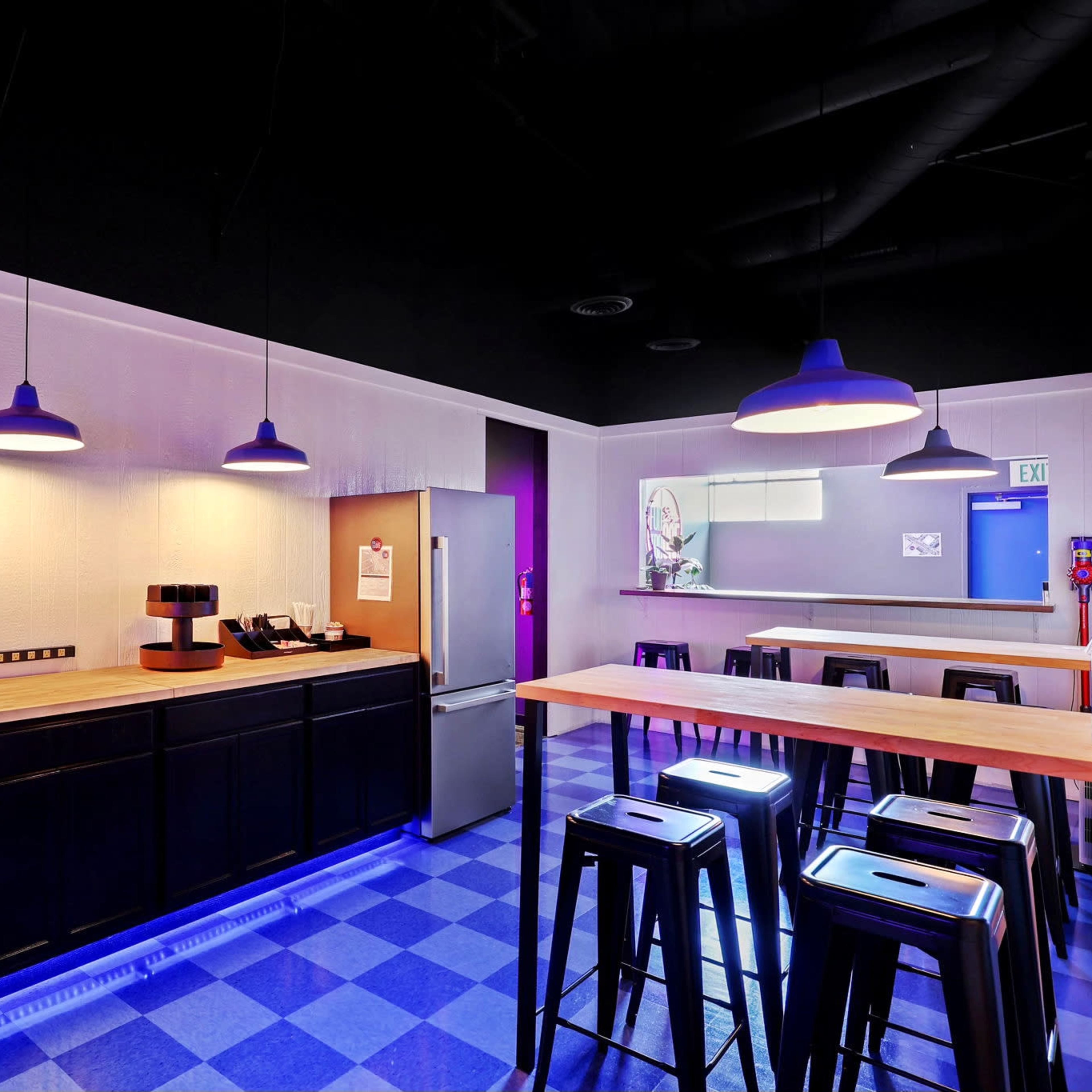 A modern kitchen space with a long wooden countertop, black bar stools, and overhead pendant lights, featuring a refrigerator and various kitchen appliances.