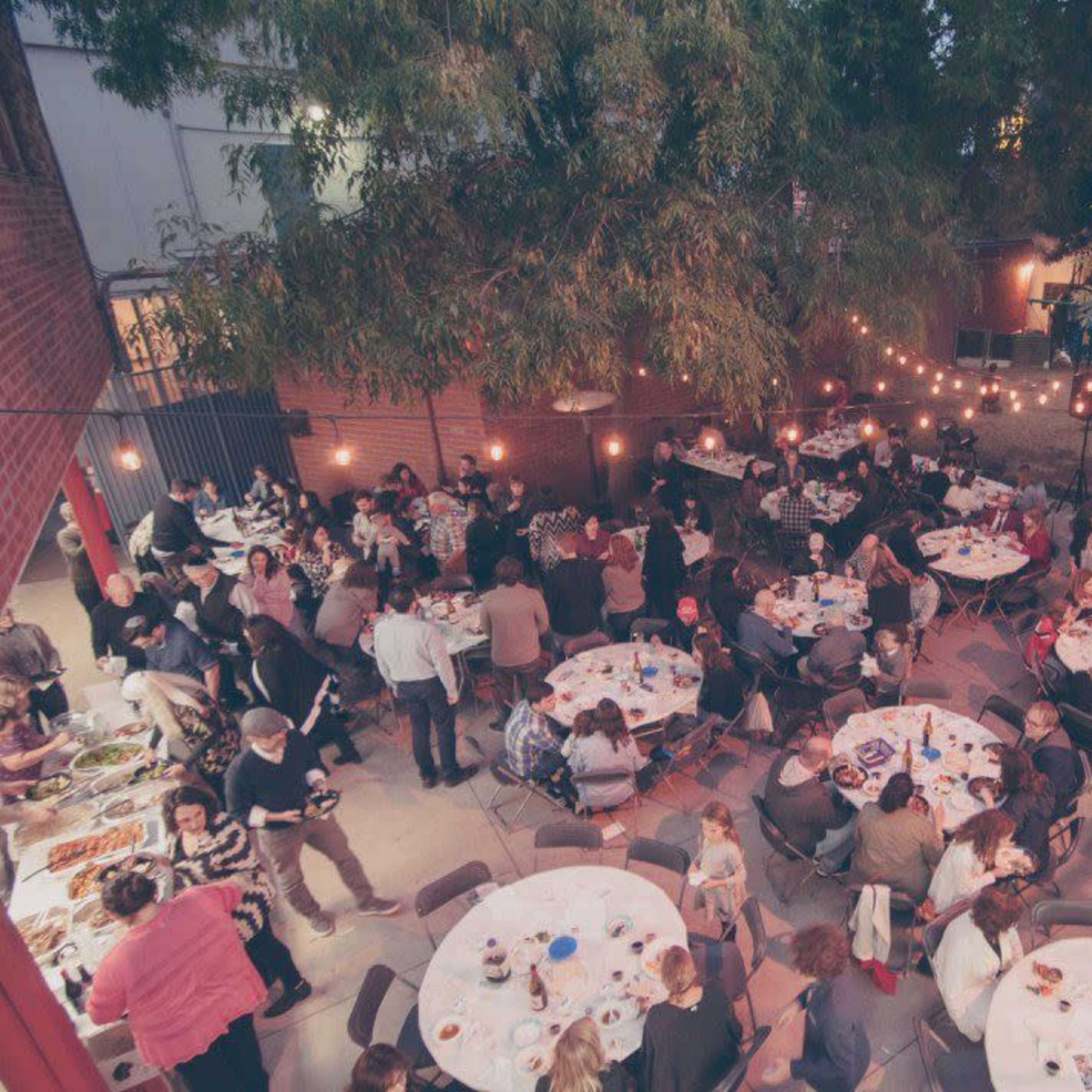 A large outdoor dining area is filled with numerous round tables, where many guests are engaged in conversation under strings of hanging lights and trees.