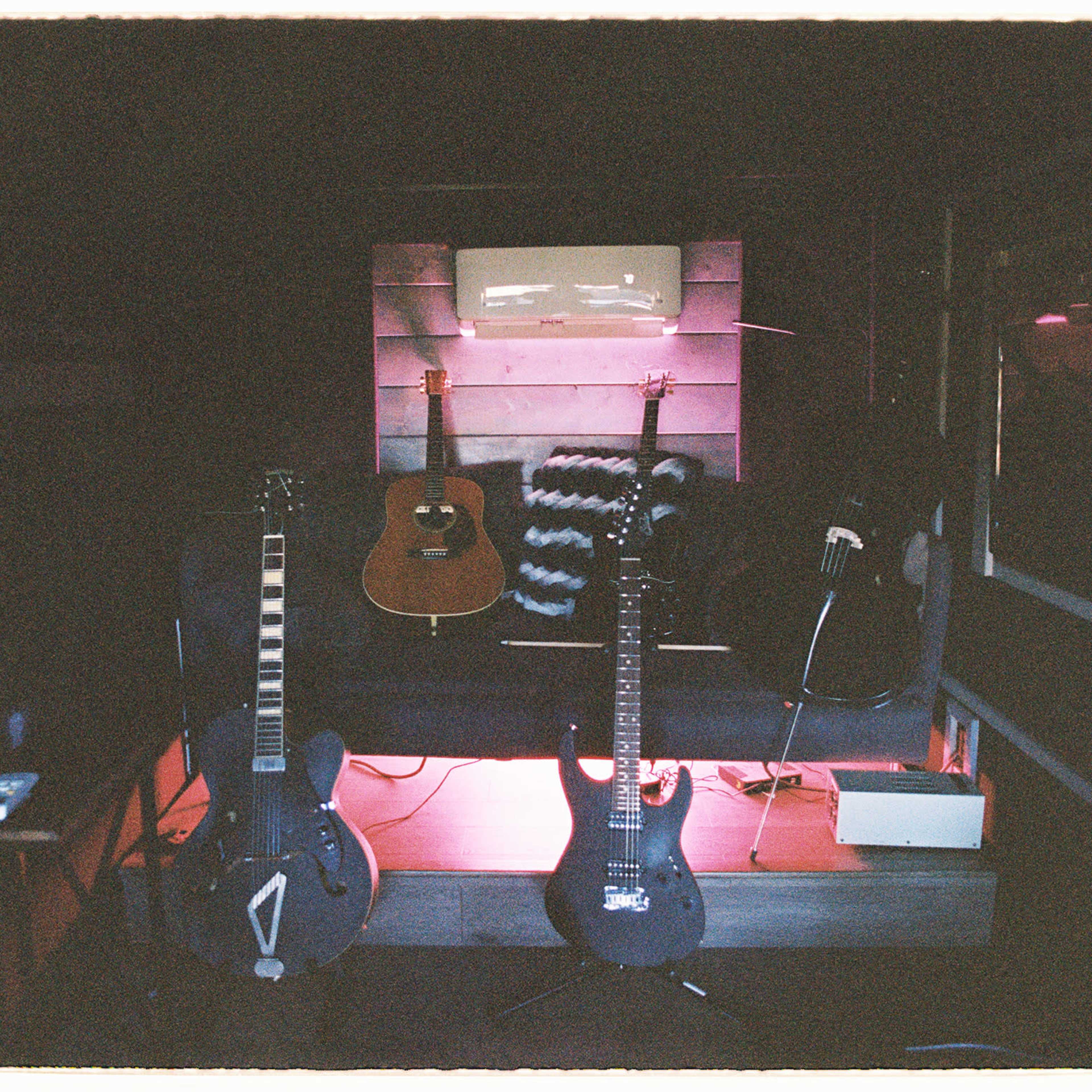 A dimly lit room featuring three guitars displayed on a shelf, with a small amplifier and a remote control nearby.