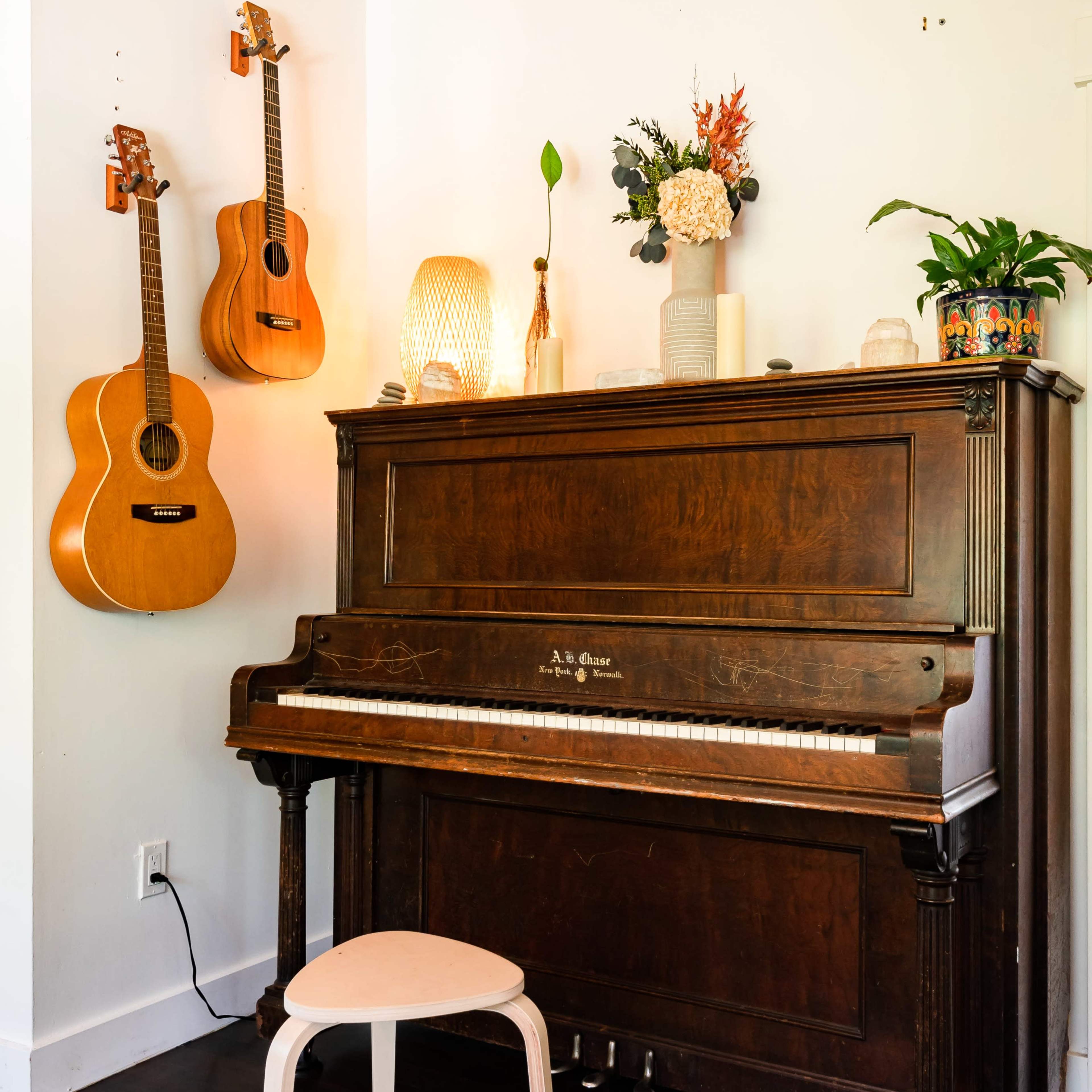 A dark wooden piano is situated against a white wall, accompanied by two guitars hung above it and a small round stool in front.