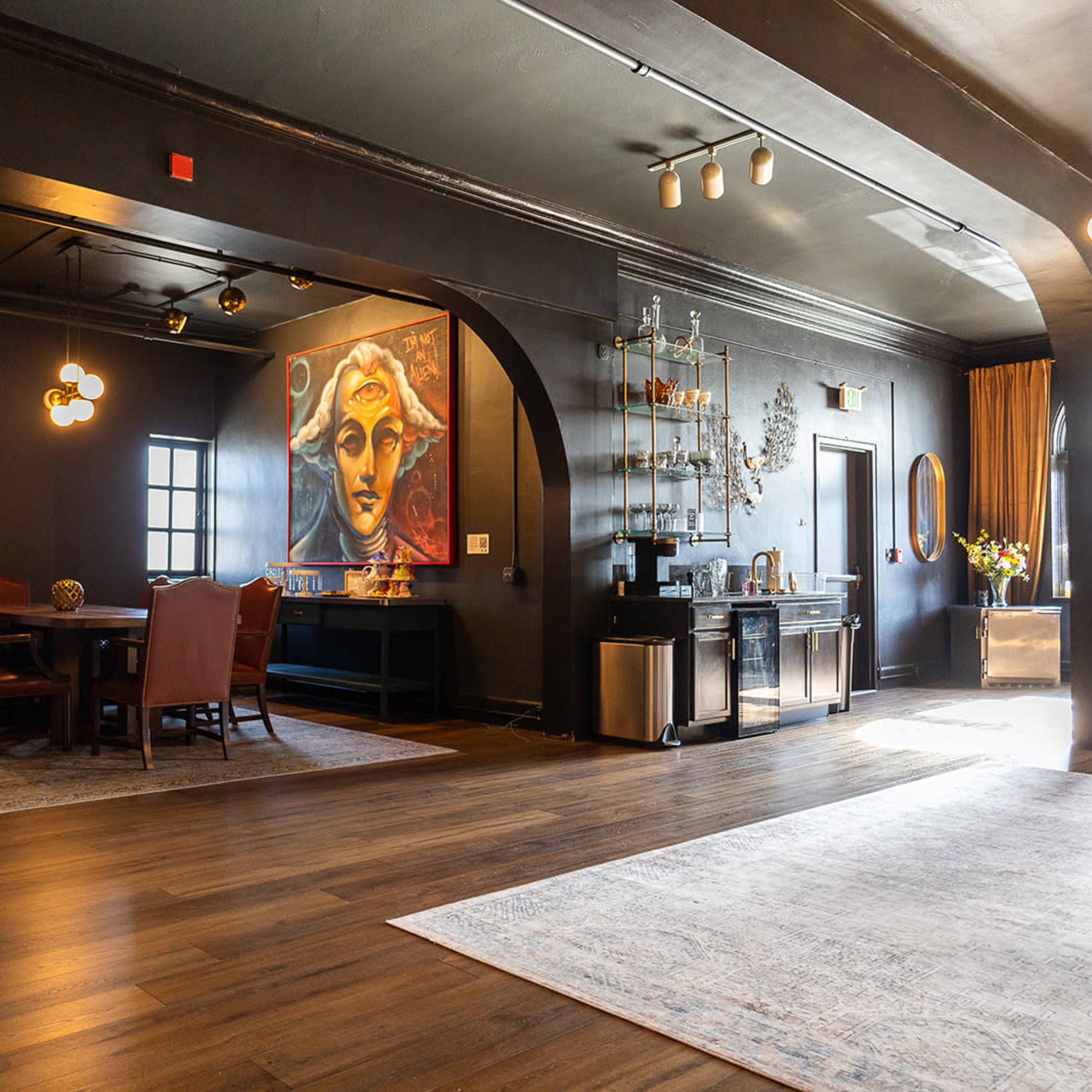 A spacious room with dark walls, a large mural of a face, a dining area with a wooden table and chairs, and a sideboard with decorative items.