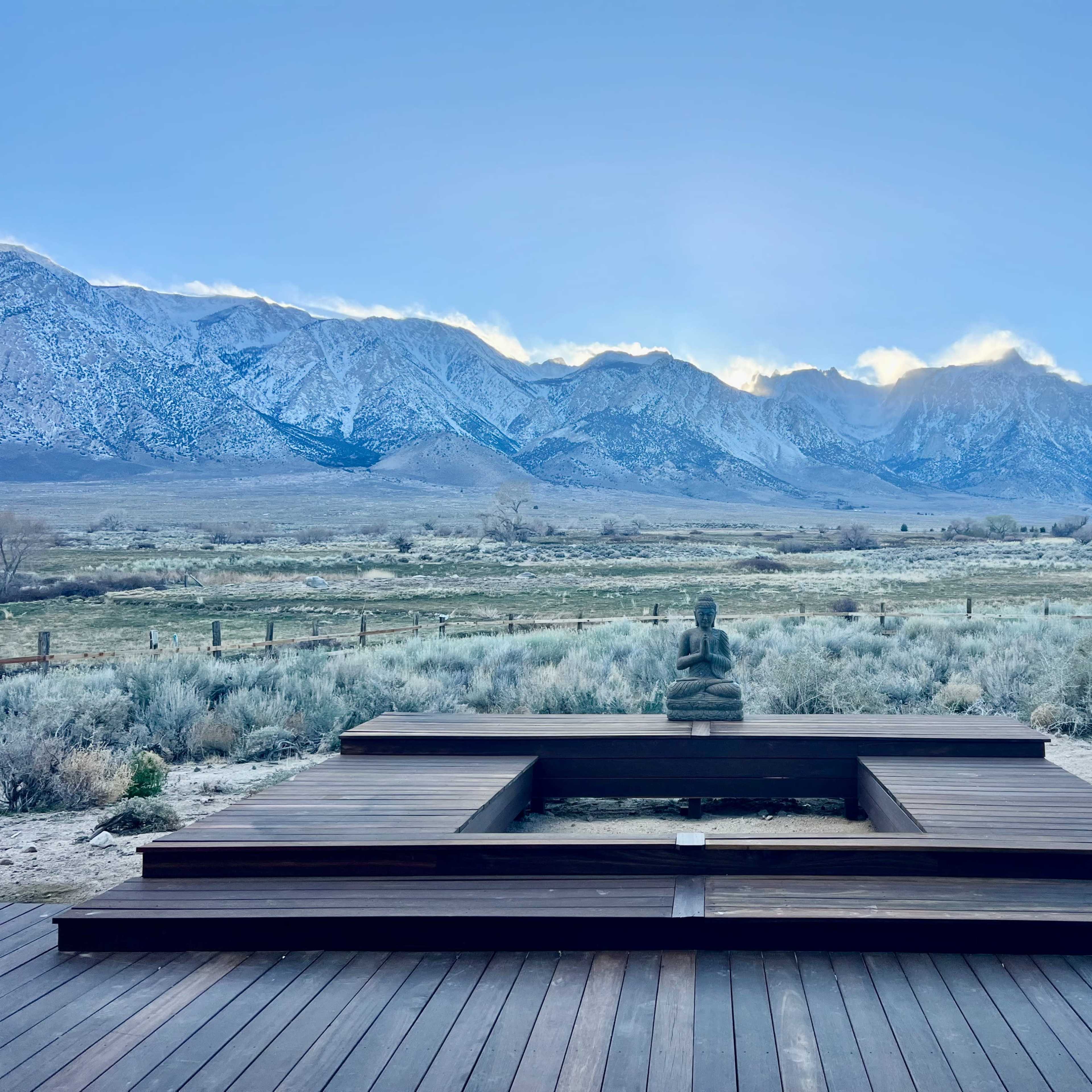 A wooden outdoor deck features a seated Buddha statue with a backdrop of snow-capped mountains and a clear sky.