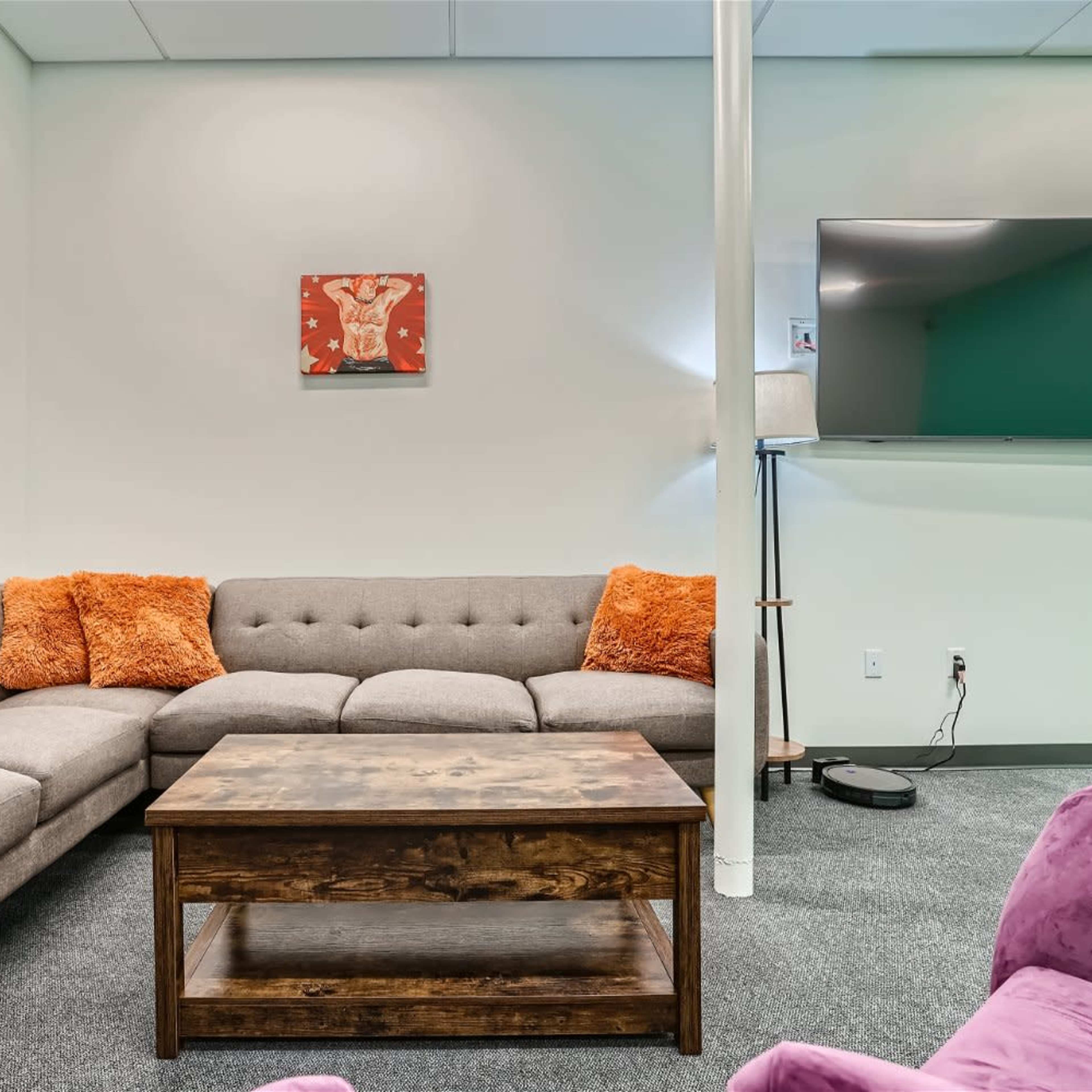 A modern lounge area with a gray sectional sofa featuring orange cushions, a wooden coffee table, and a mounted television on the wall.
