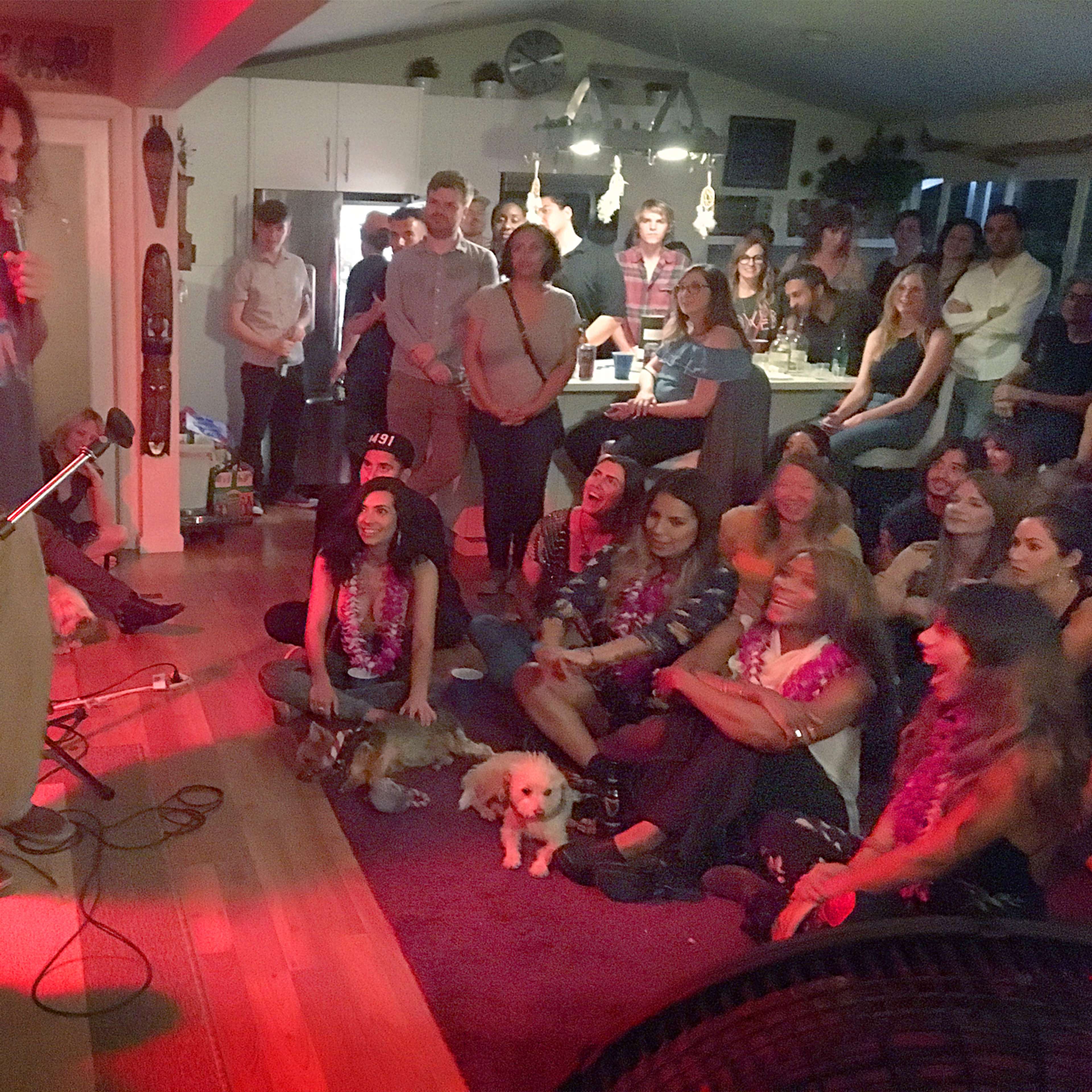 A crowd gathers in a living room, attentively watching two speakers on a small stage while dogs sit among the audience.