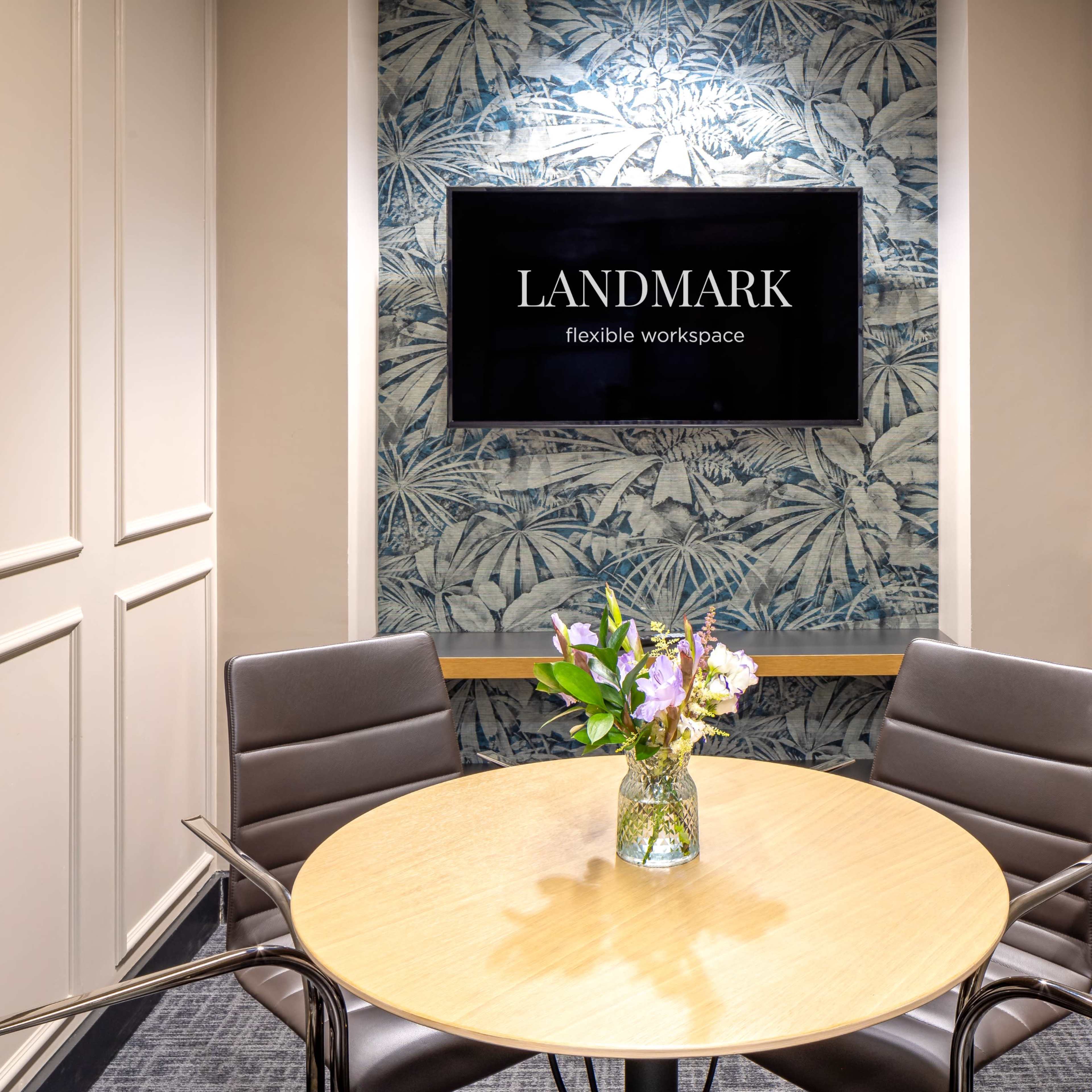 A small conference room features a round wooden table surrounded by four black chairs, with a floral arrangement in the center and a large screen displaying the "LANDMARK" logo on the wall.