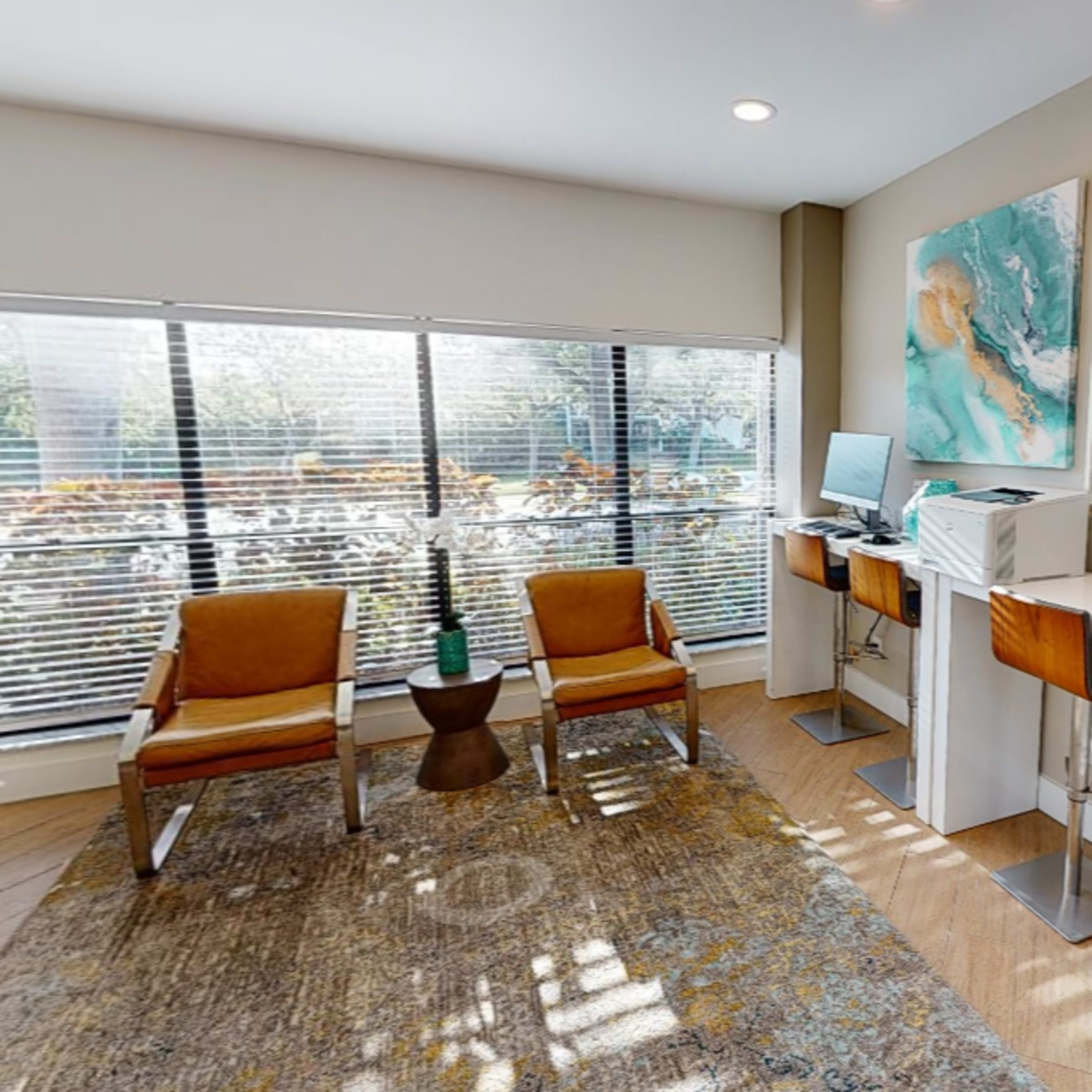 A modern office space with two tall desks, a computer setup, orange armchairs, and large windows letting in natural light.