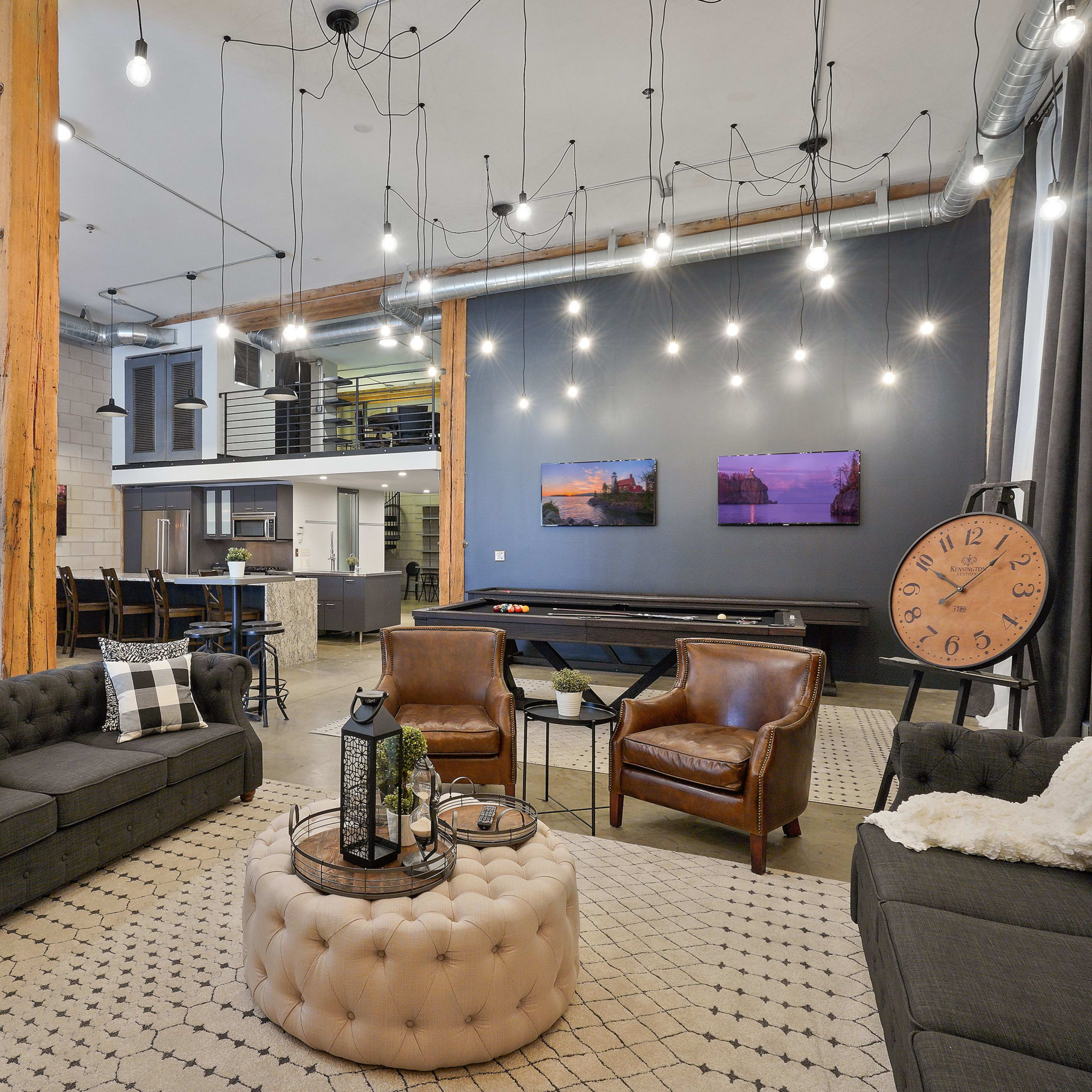 A modern open-concept living space with a combination of seating areas, a dining area, and a kitchen, featuring industrial-style decor and hanging light fixtures.