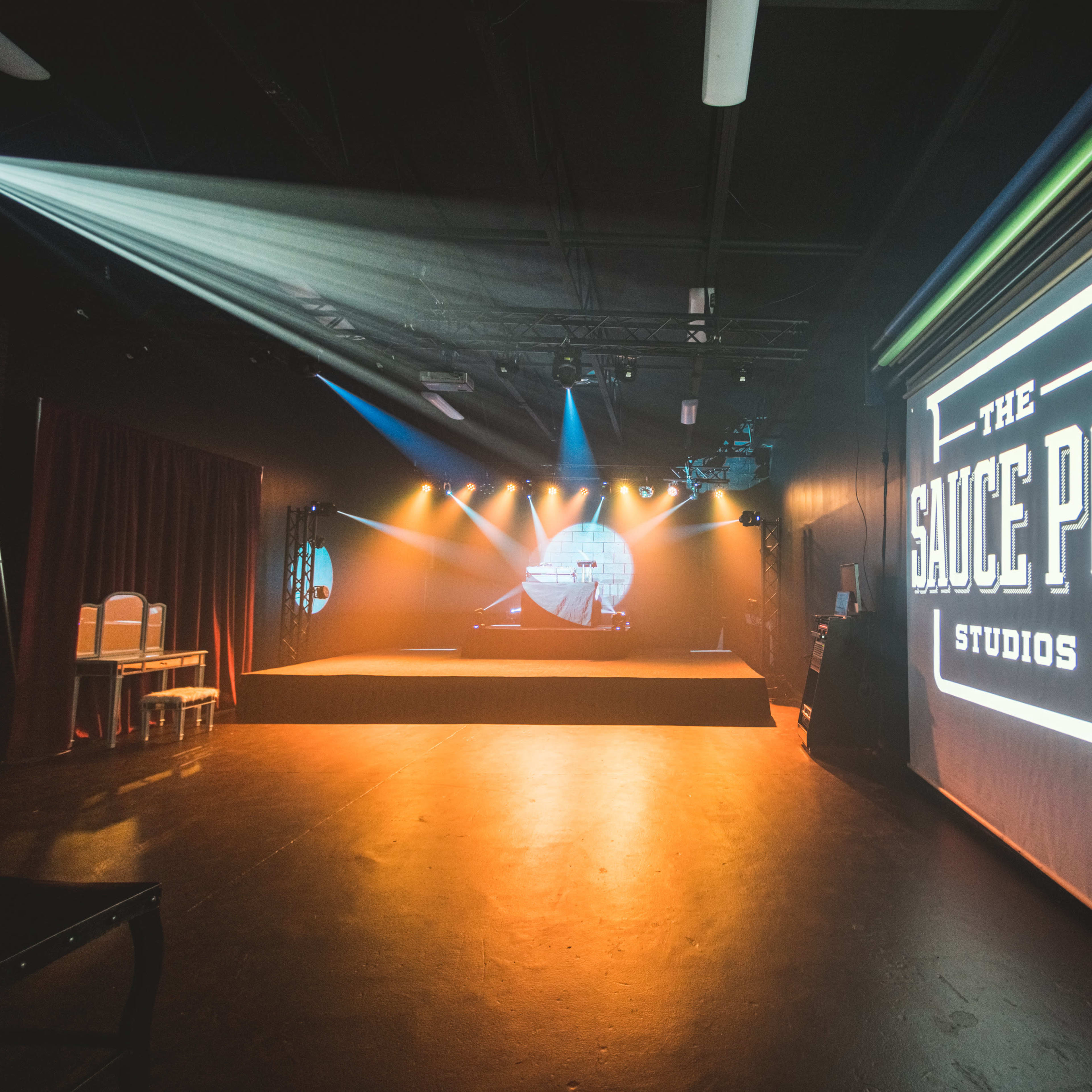 An interior of a performance studio called "The Sauce Pot Studios," featuring a stage with lighting and a large logo displayed on a screen.