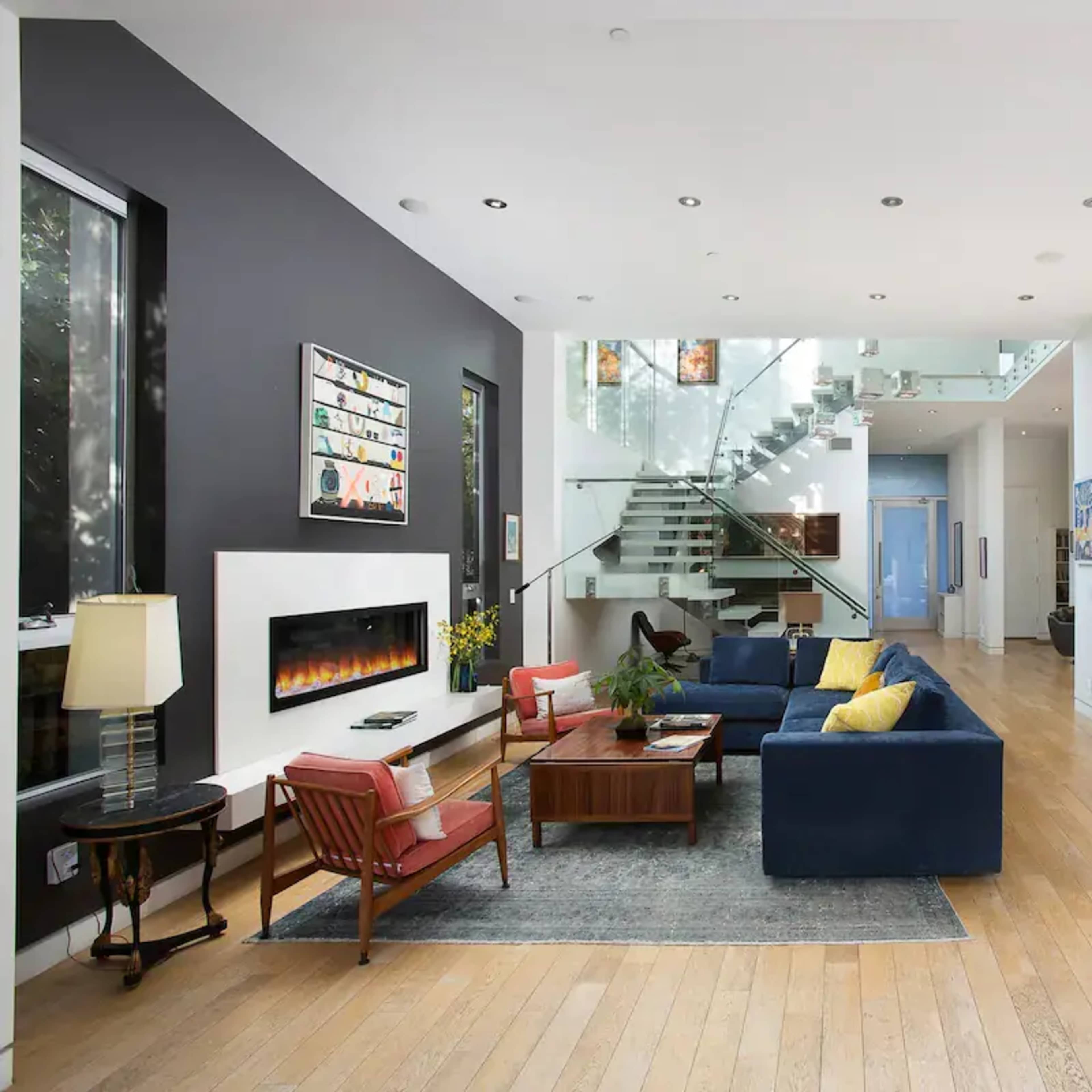 A modern open-concept living area with a sleek staircase, large windows, and a fireplace, featuring a sectional sofa, wooden dining table, and various furnishings.