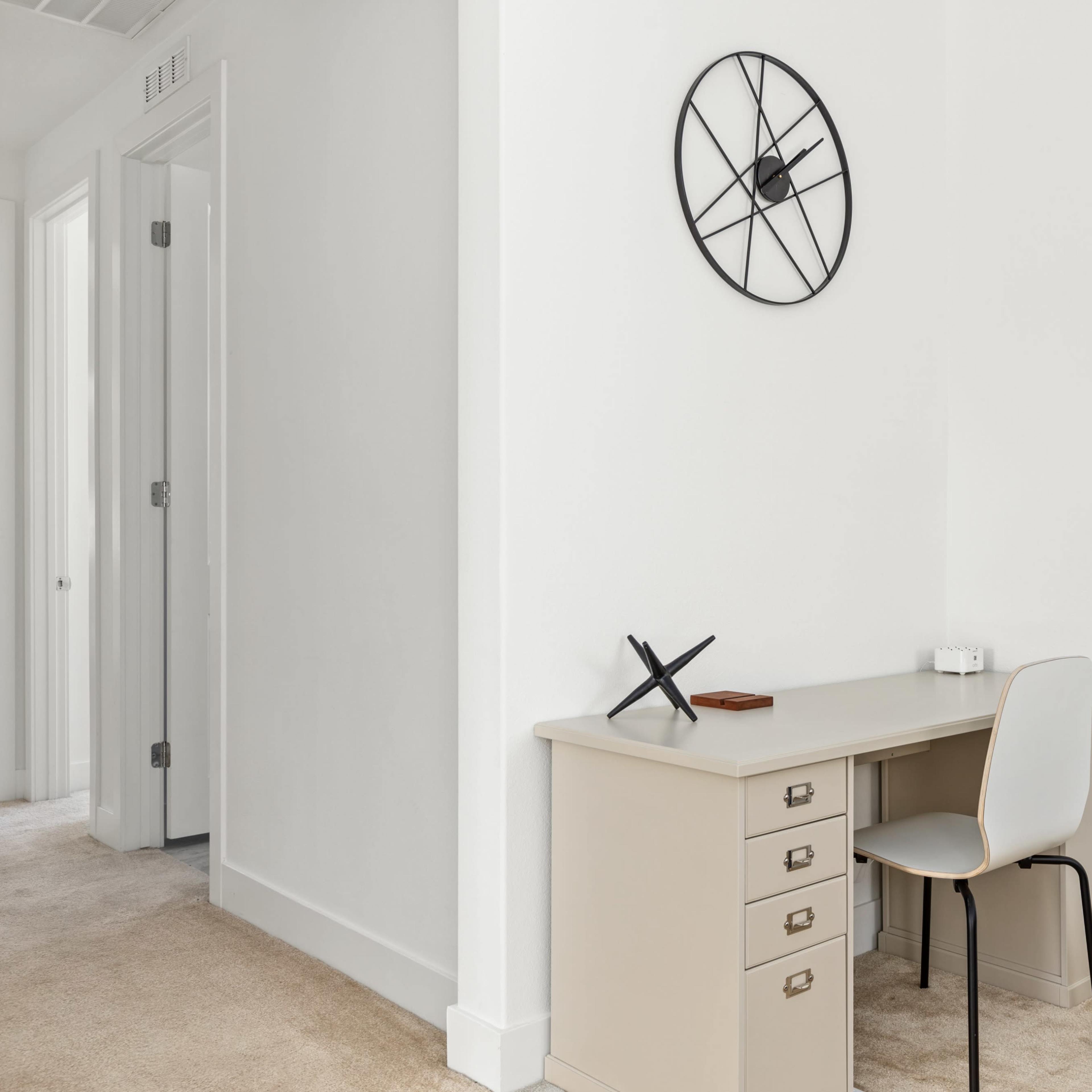 A minimalist workspace features a light-colored desk with a chair and a wall clock in a room with white walls and carpet.