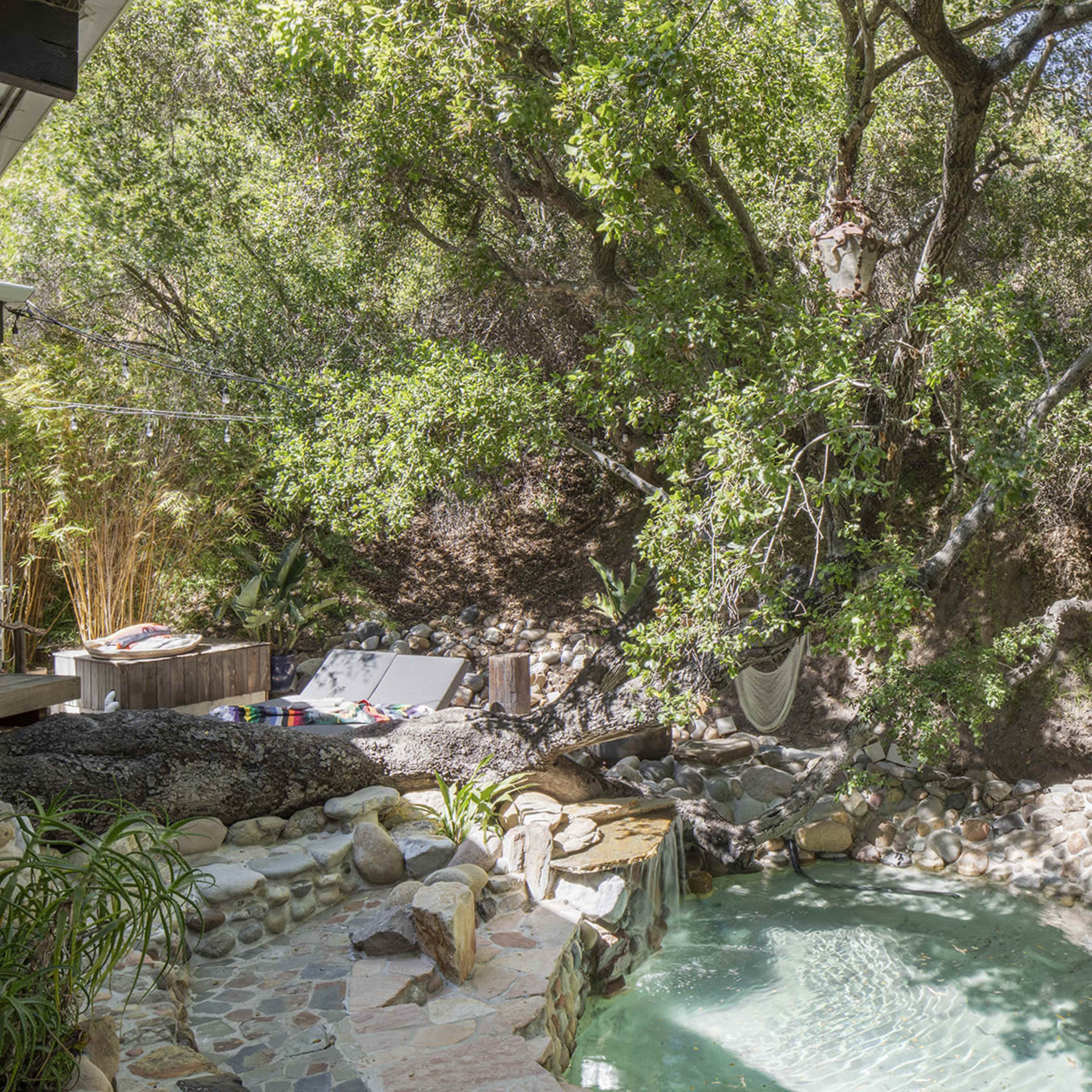 A small, natural swimming pool surrounded by rocks and lush greenery, with lounge chairs and a hammock nearby.