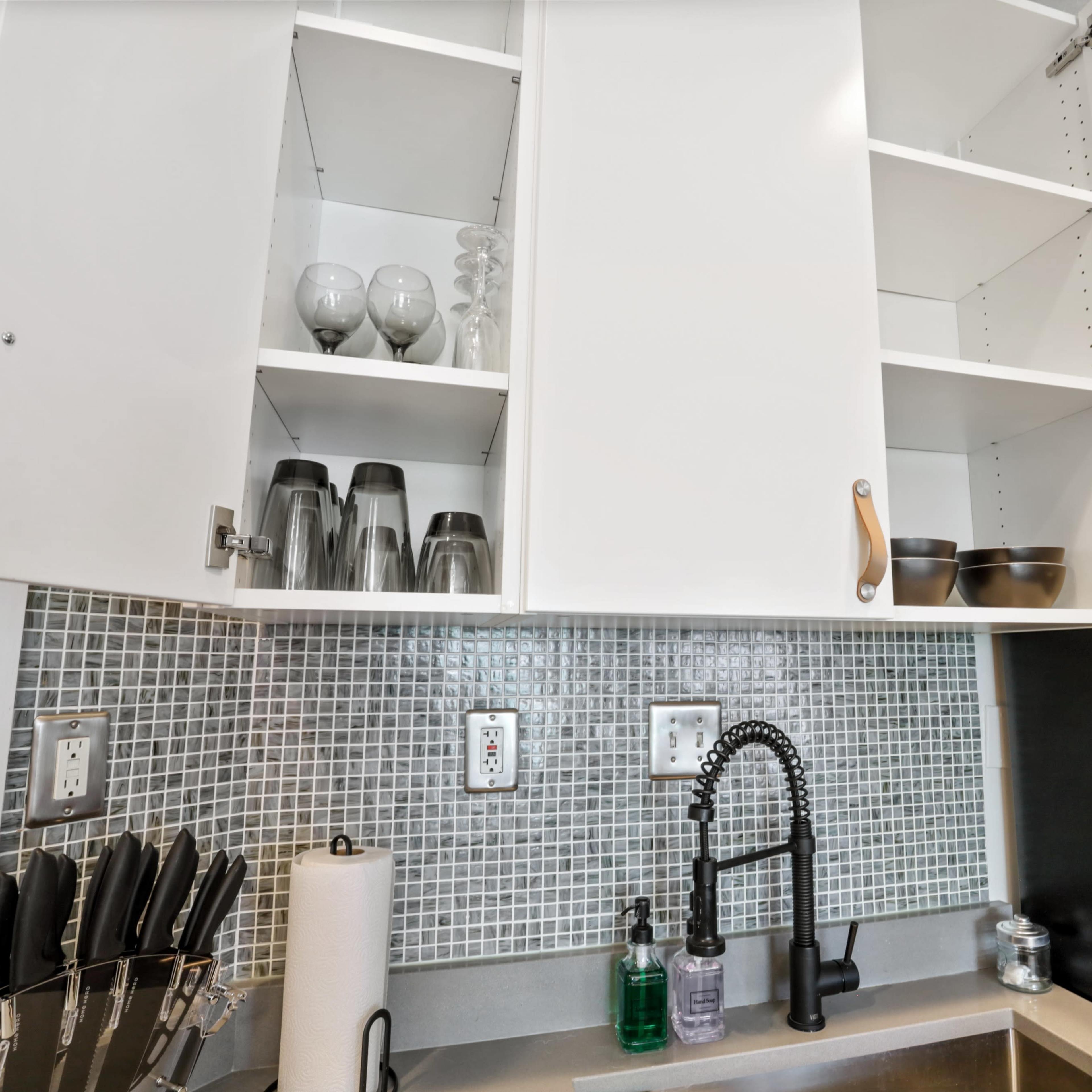 A kitchen with open cabinets displaying glassware and dishes, a modern sink with a black faucet, and a backsplash of mosaic tiles.