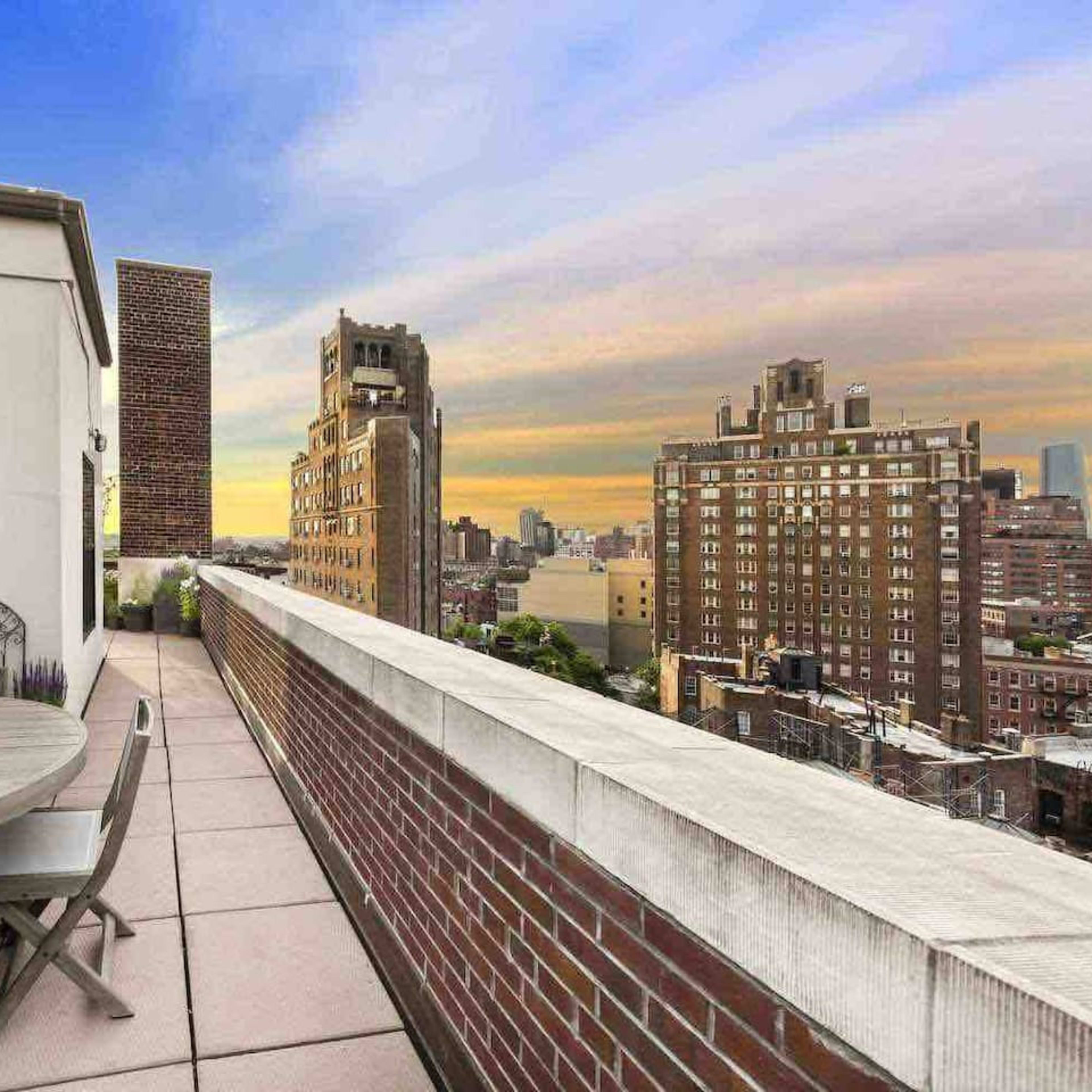 A rooftop terrace features a round table with sunflowers and a view of city buildings against a sunset sky.