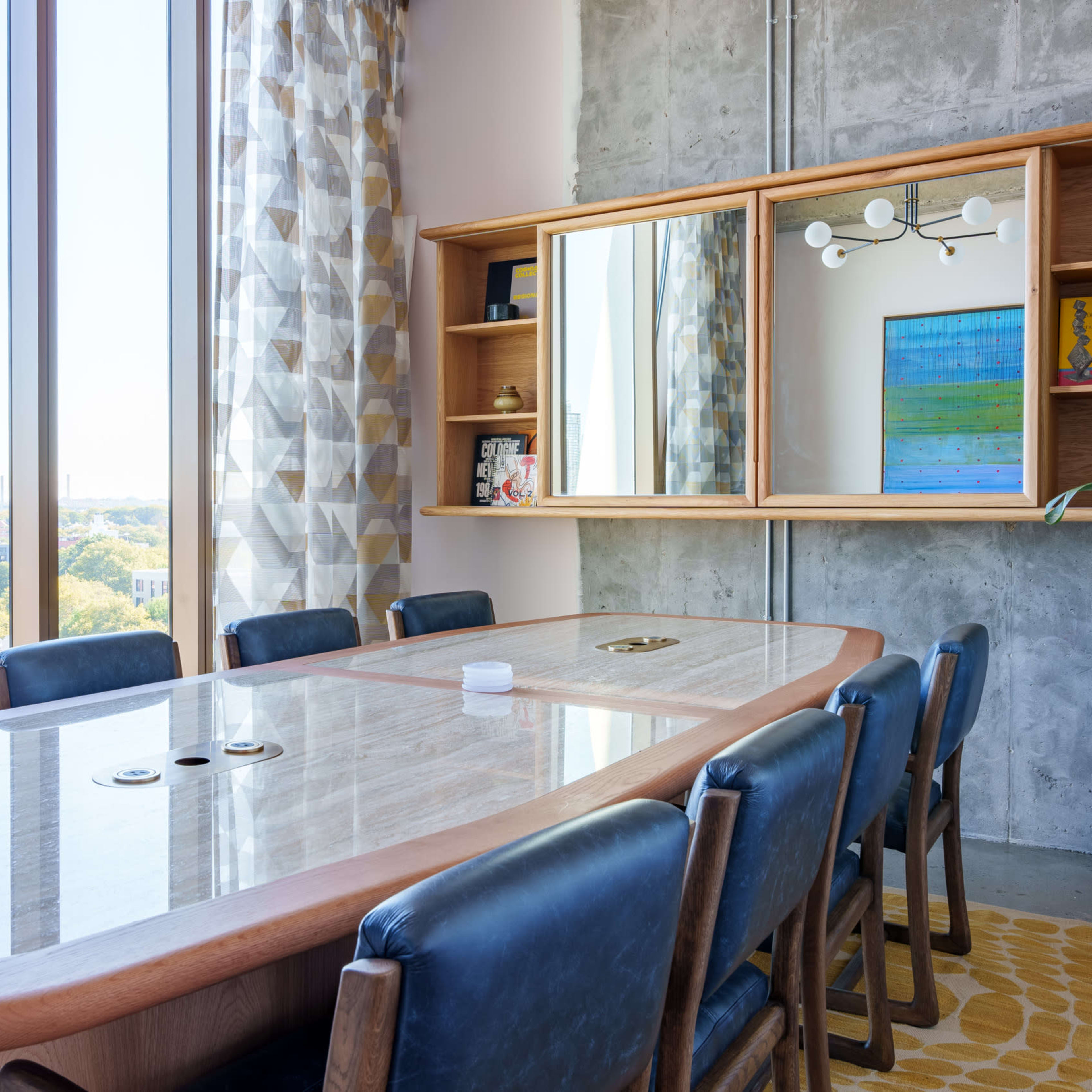 A modern conference room features a large wooden table surrounded by blue chairs, with a mirror and decorative shelving against a concrete wall.