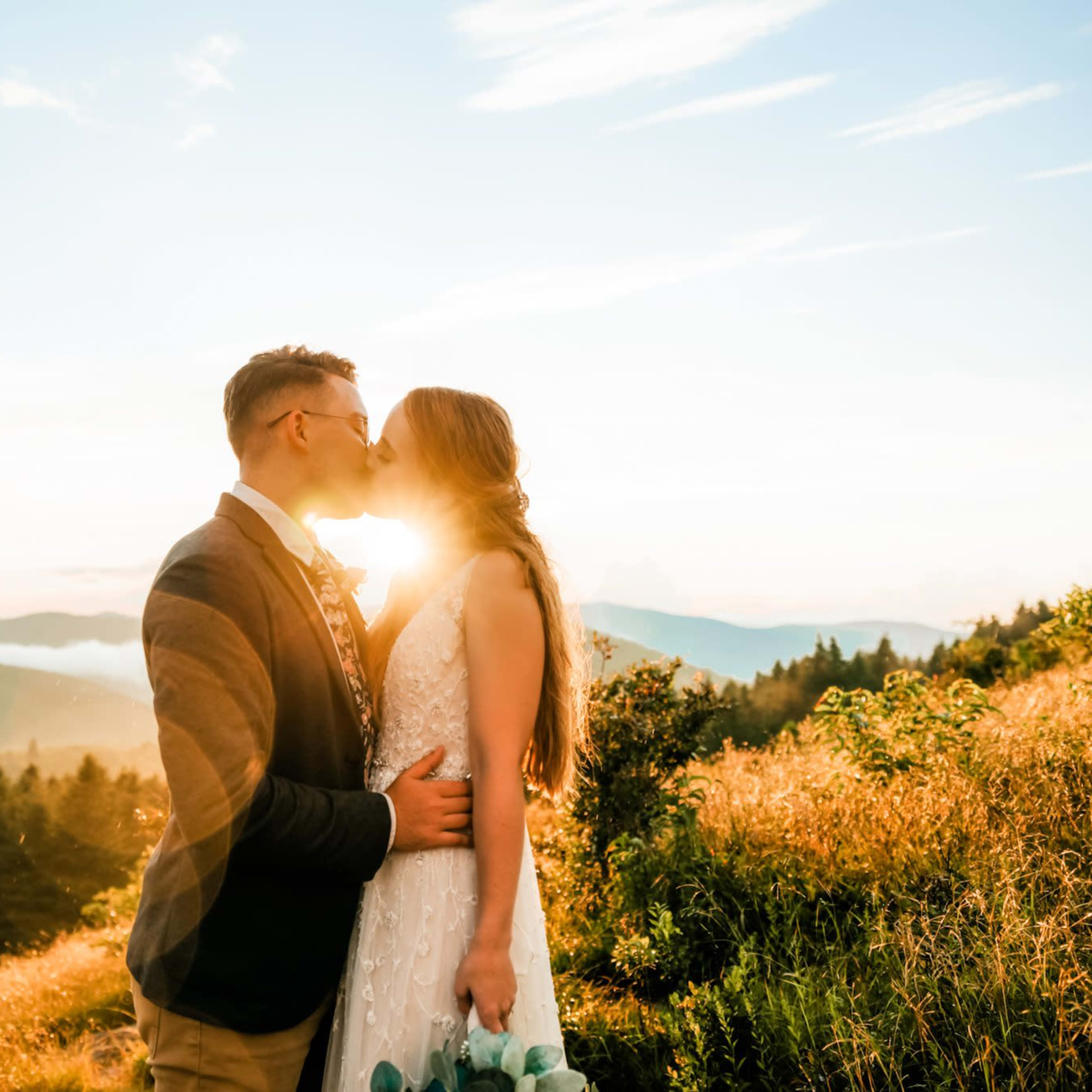 A couple stands in an open field at sunset, embracing and kissing with mountains in the background.