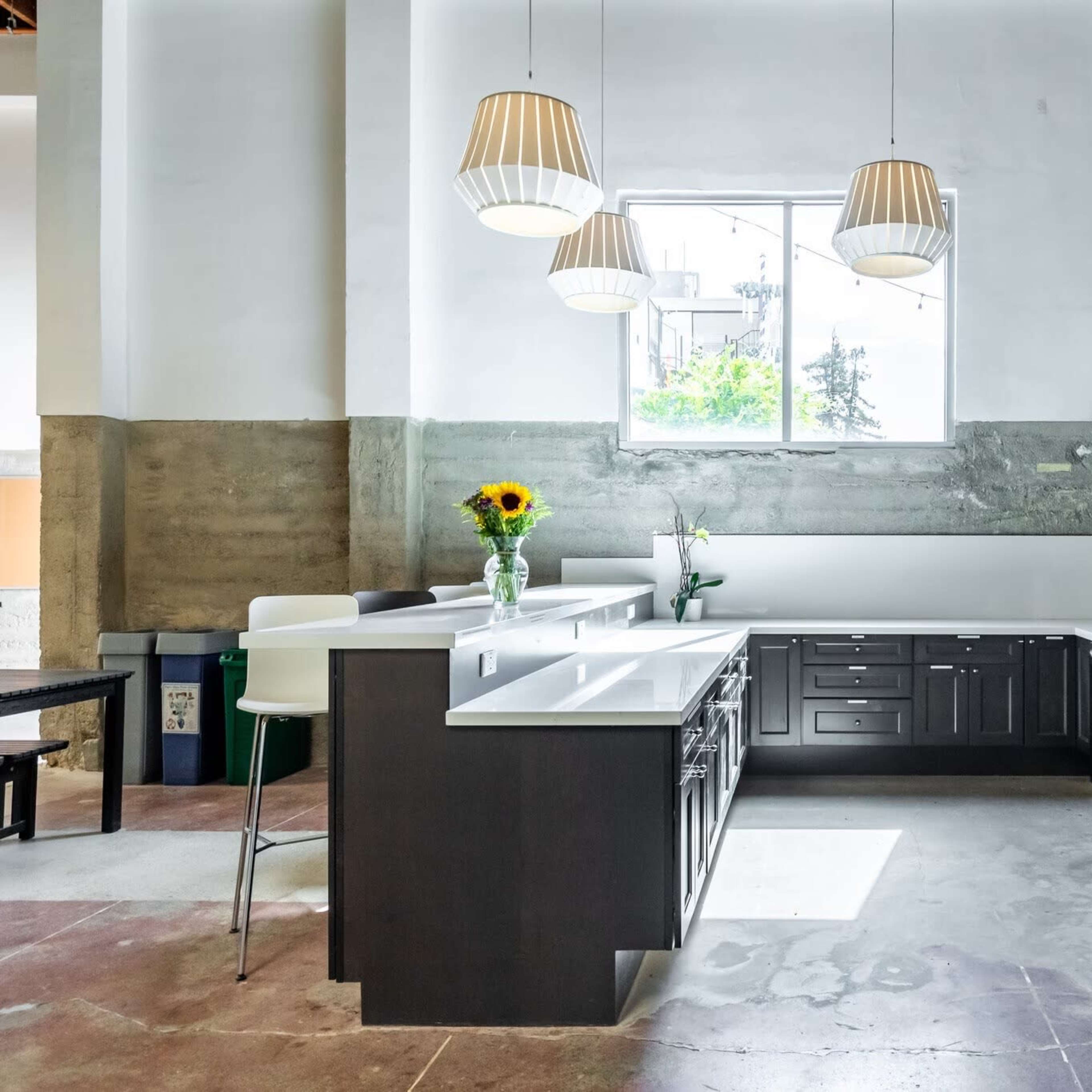 A modern kitchen with dark cabinetry and white countertops, featuring a central island and large windows that allow natural light to enter.