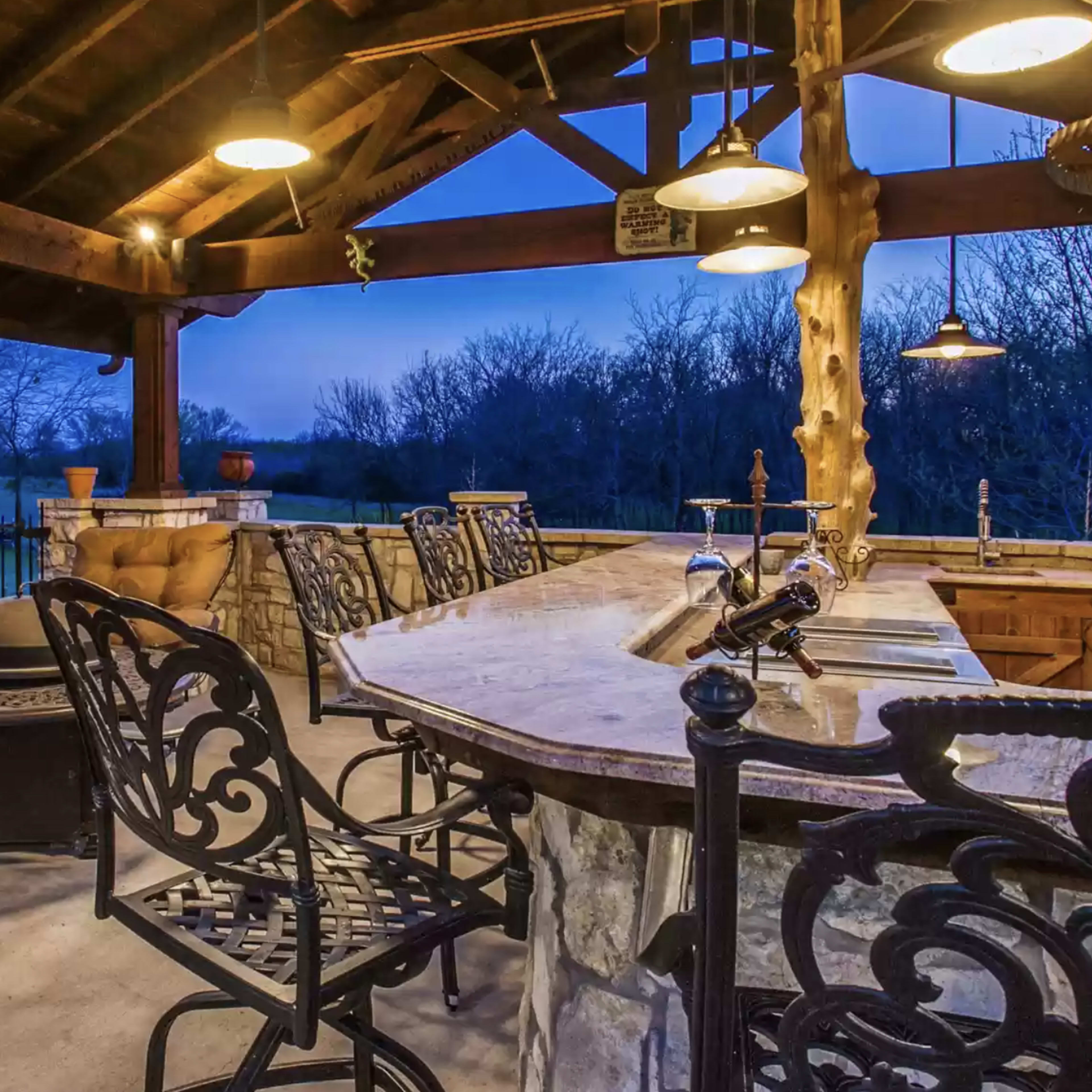 An outdoor bar area with metal chairs, a marble countertop, and a wooden structure under low lighting at twilight.