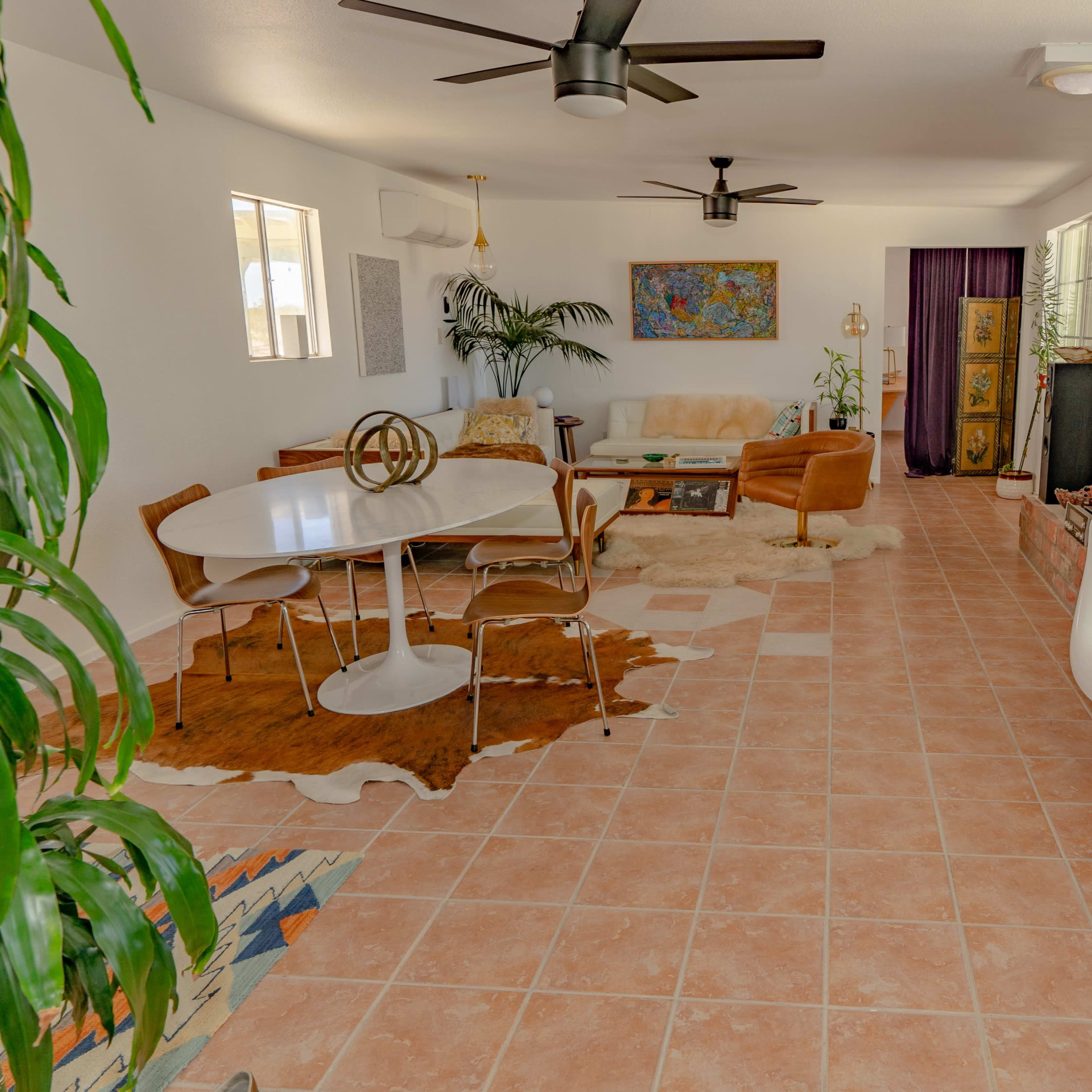A living room features tiled flooring, a circular dining table, brown seating, and large windows with greenery visible.