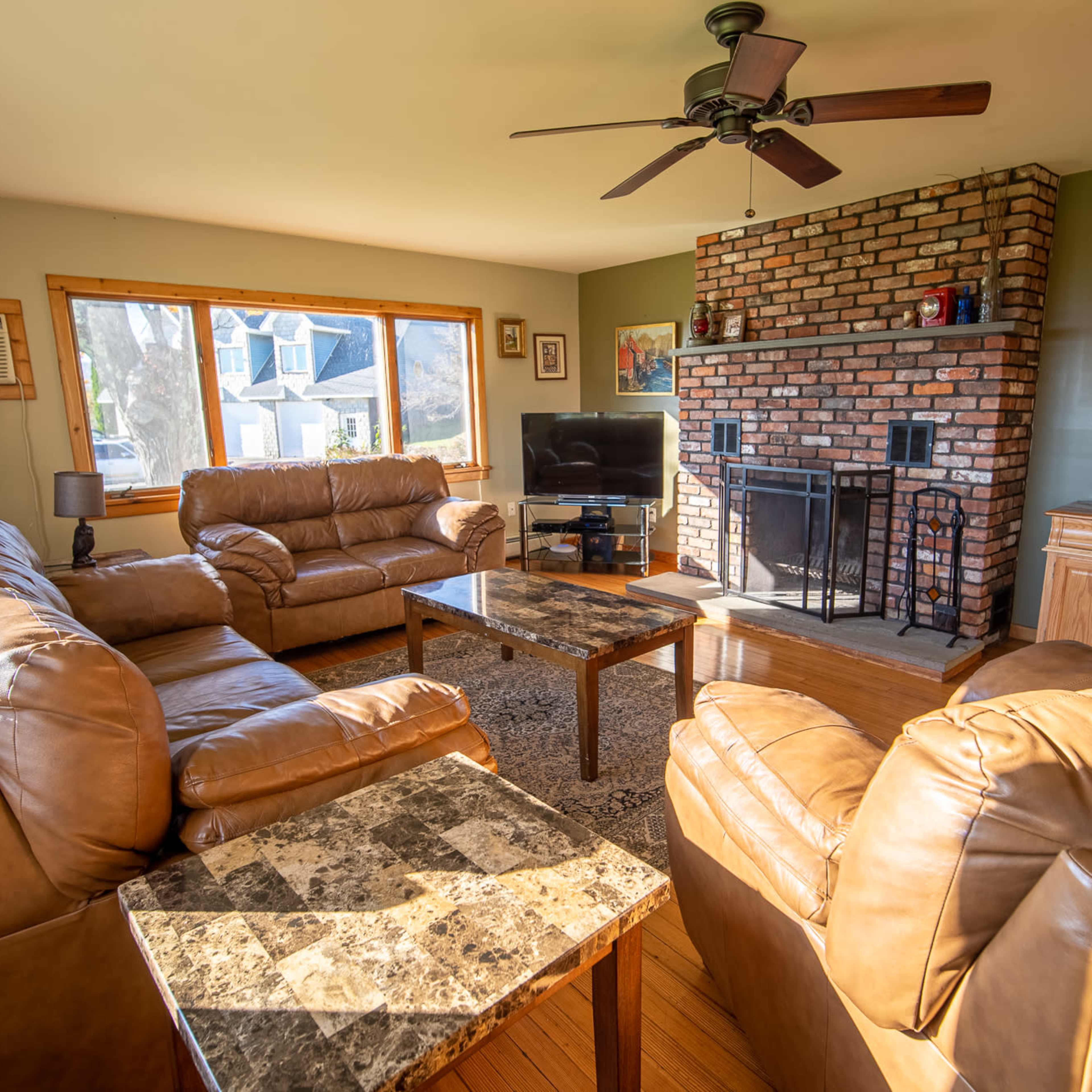 A living room furnished with leather sofas, a coffee table, and a brick fireplace, with a large window providing natural light.