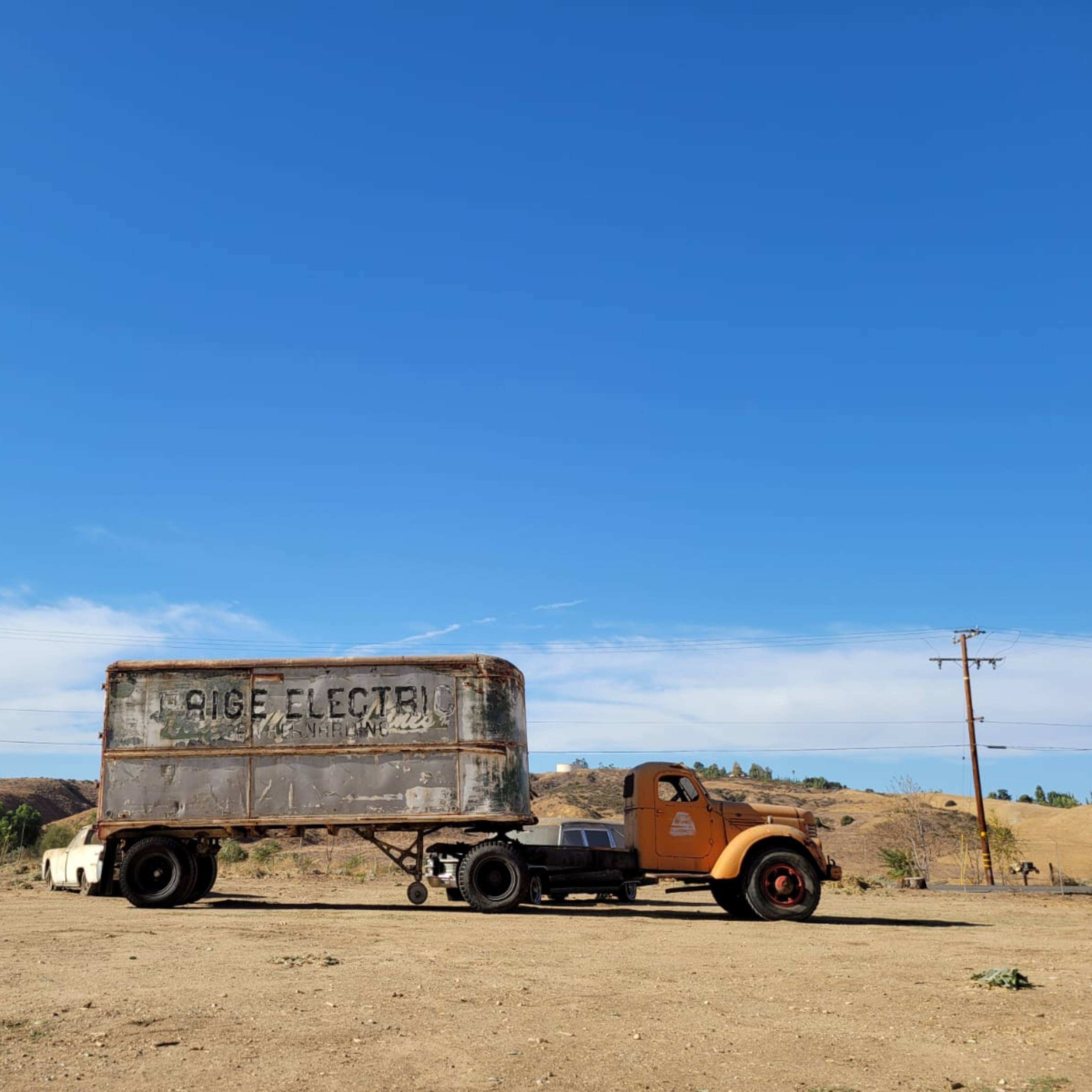 An old, rusty truck with a large trailer parked on a dirt lot under a blue sky with distant hills.