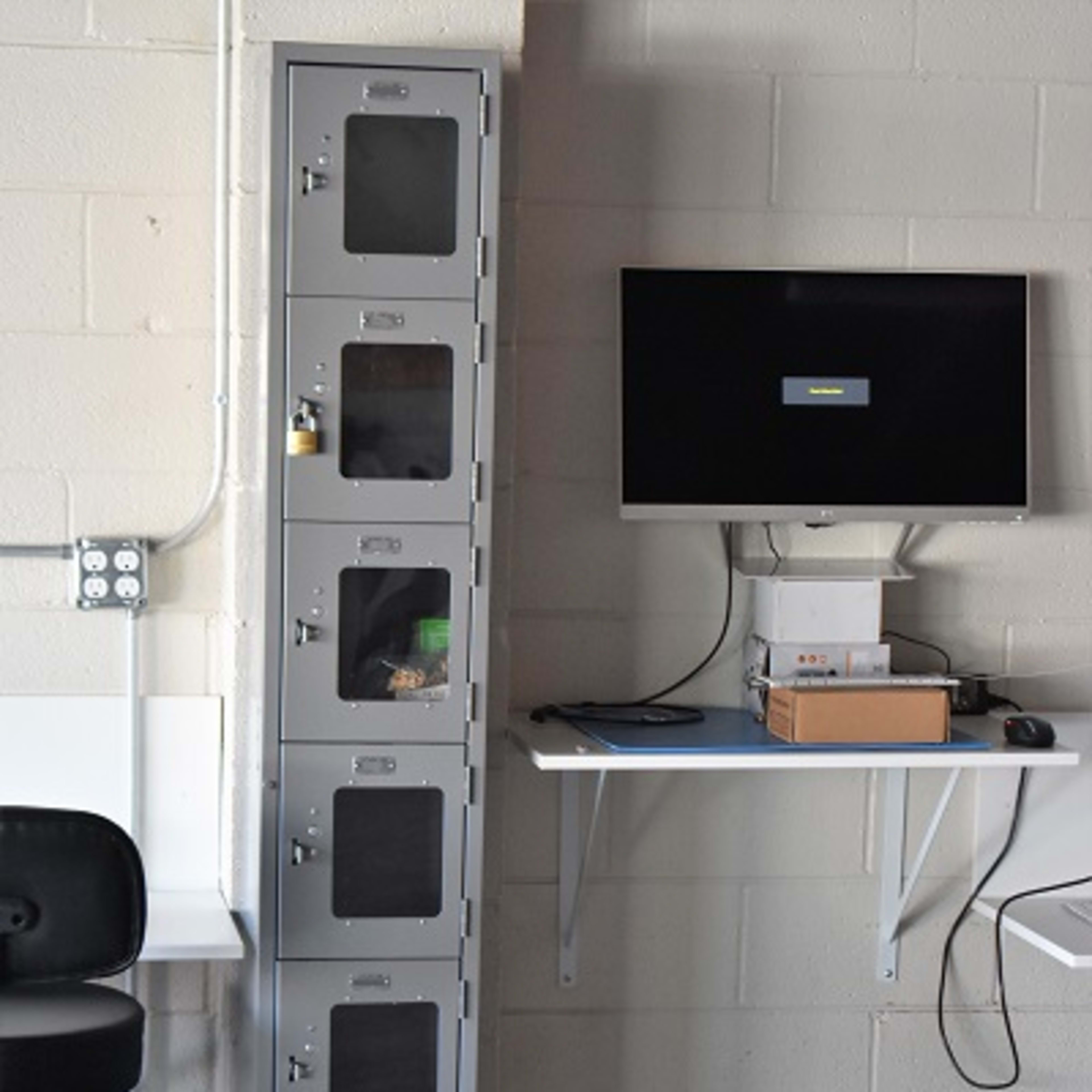 A gray metal locker unit stands against a wall with a computer monitor mounted on a shelf nearby.