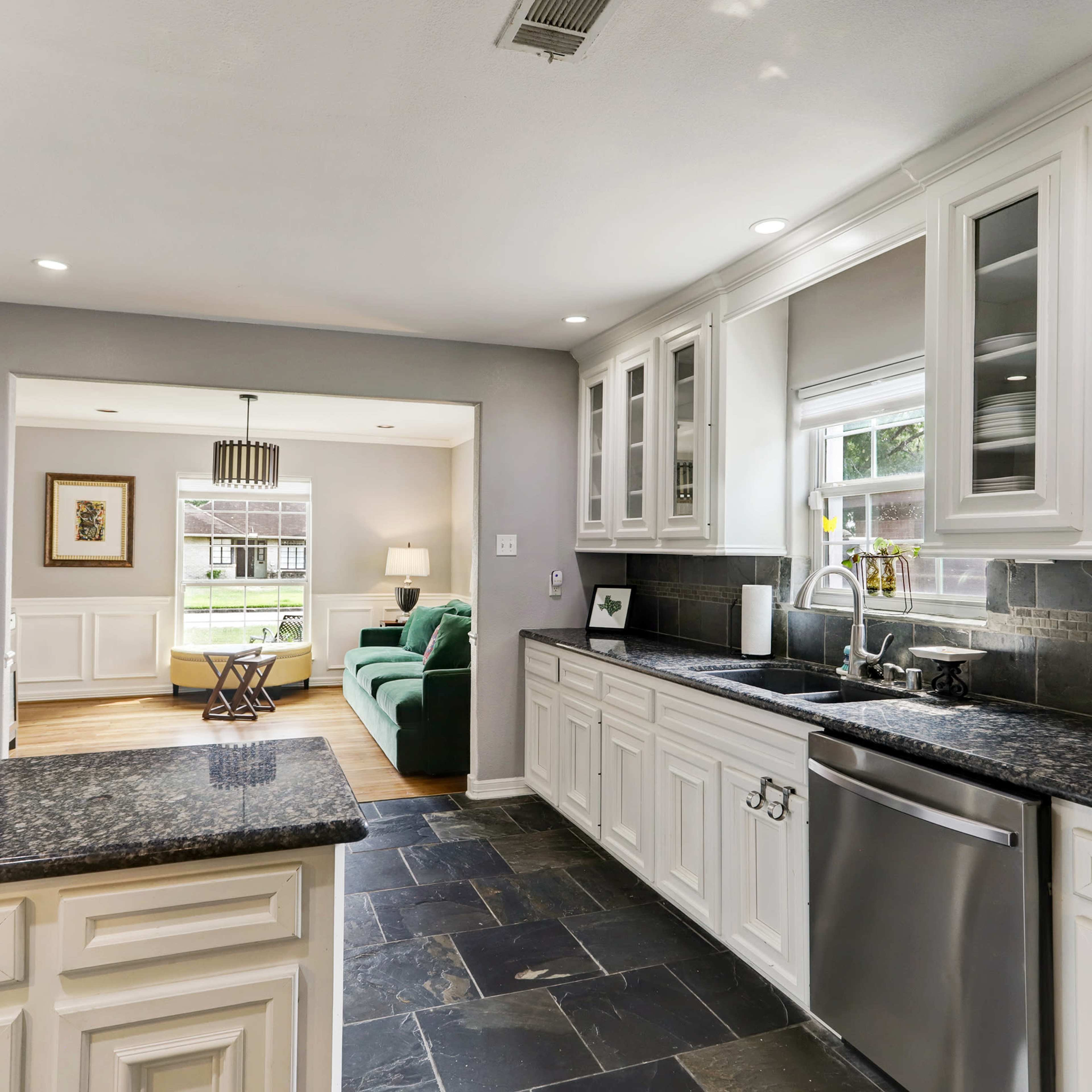 A modern kitchen with white cabinets, dark stone countertops, and stainless steel appliances, connected to a living area with a green sofa.