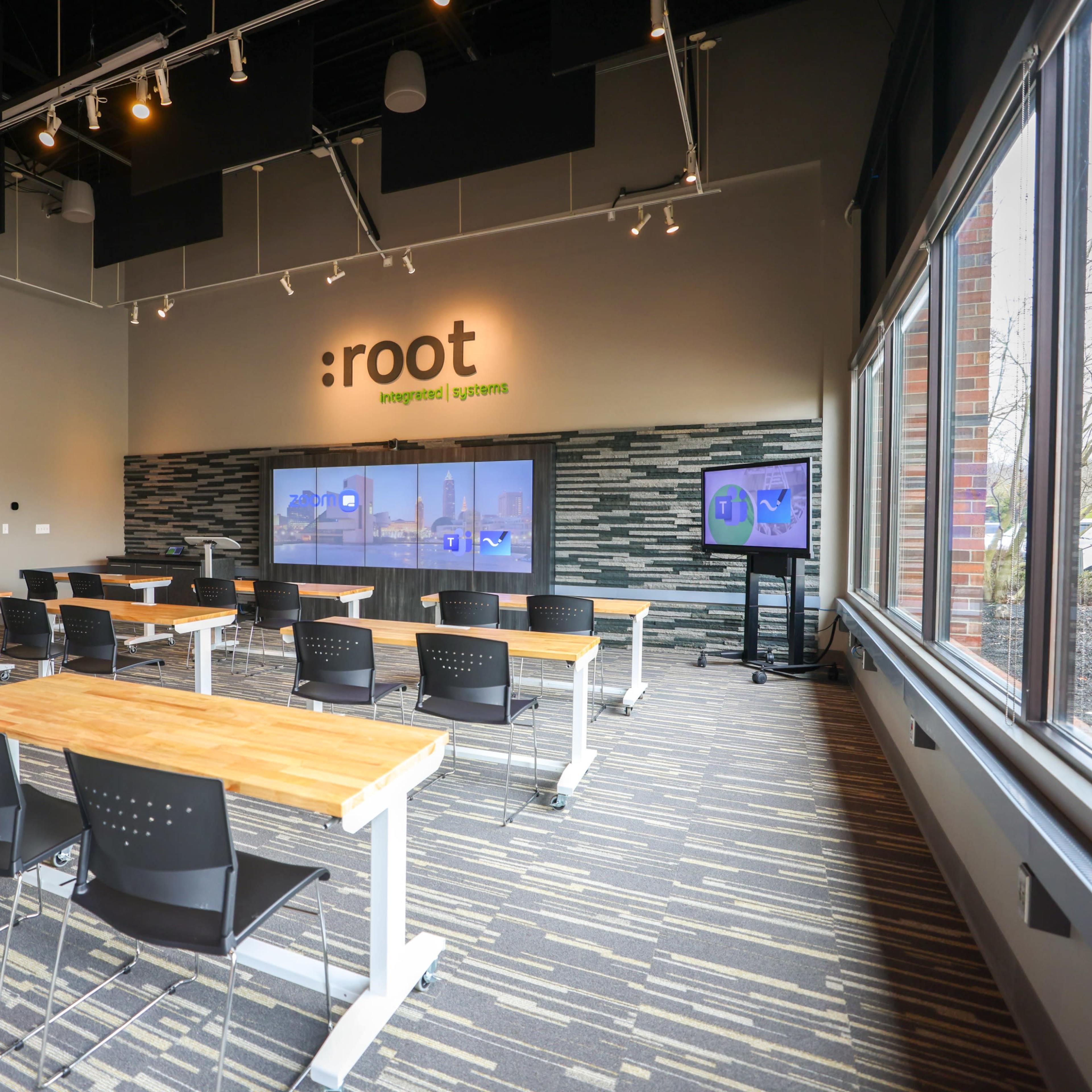 A modern classroom with multiple wooden tables and black chairs arranged facing a large screen on the wall.